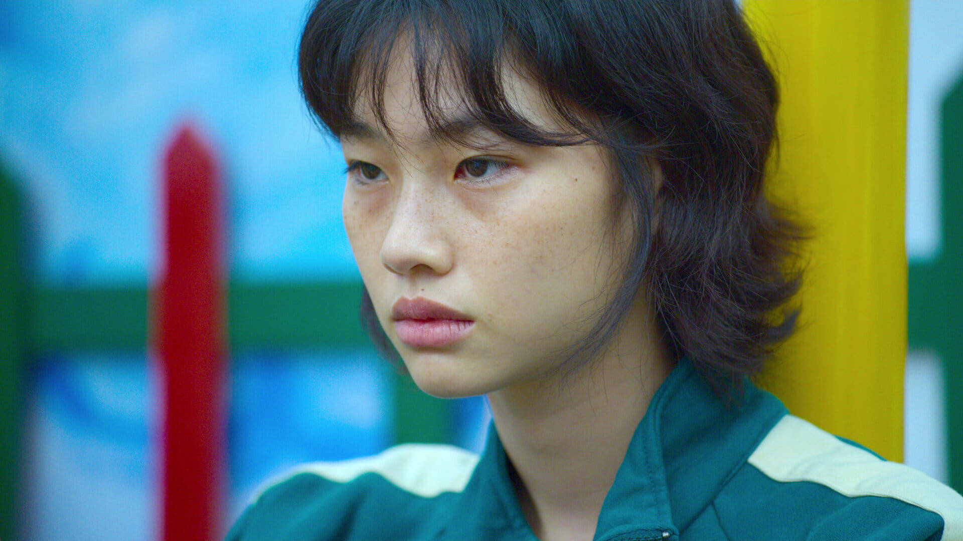 Hoyeon 桌布 壁紙 — 一位深色頭髮、有雀斑的年輕女性在色彩繽紛、模糊的背景中若有所思地凝視。