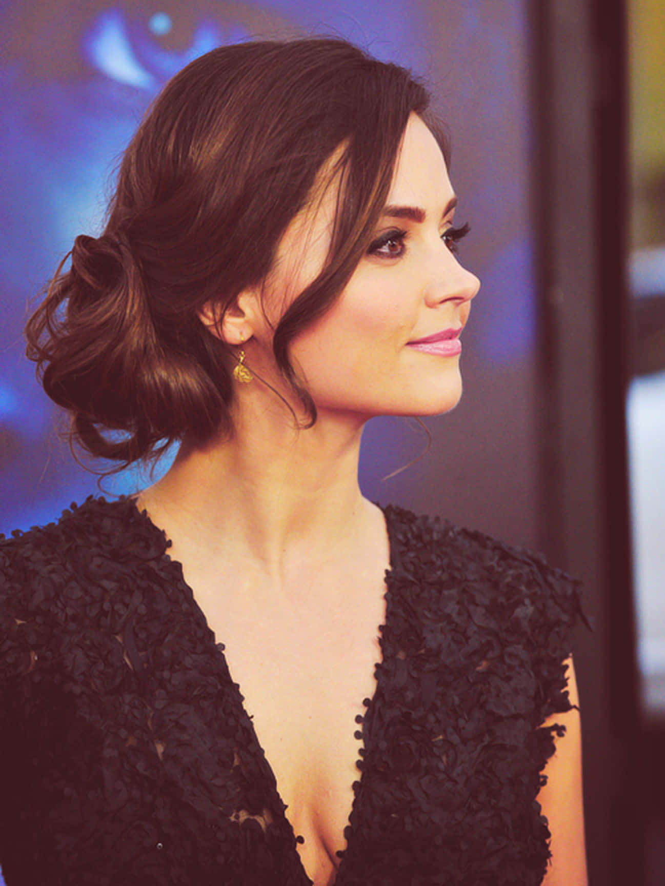 Jenna Coleman 桌布 壁紙 — 一位深色頭髮、梳著優雅盤髮的女性，在模糊的藍色背景前穿著黑色紋理連衣裙和金色耳環。