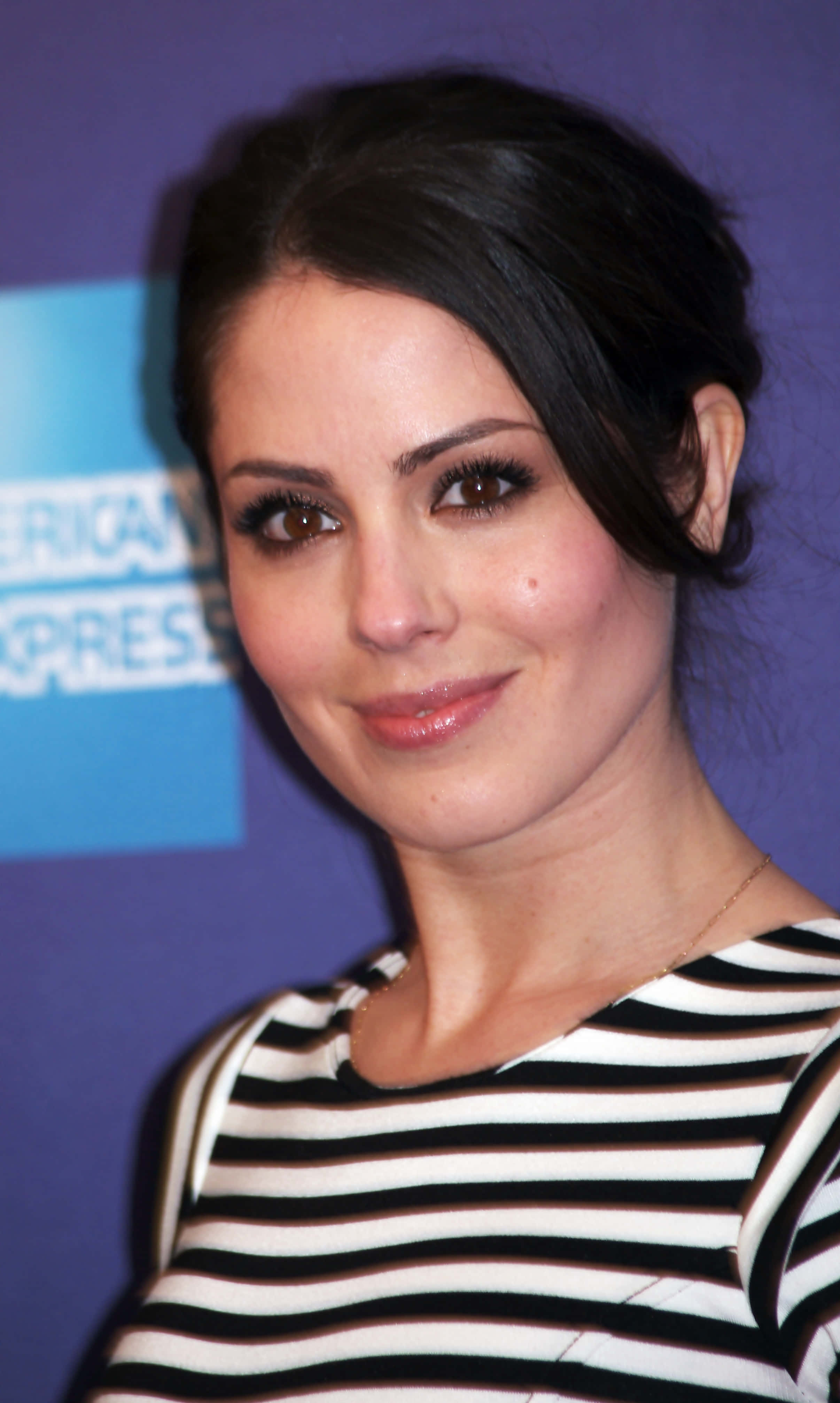 Michelle Borth 桌布 壁紙 — 一位深色頭髮、濃密睫毛的女性，穿著黑白條紋上衣，在藍色品牌背景前溫柔微笑。