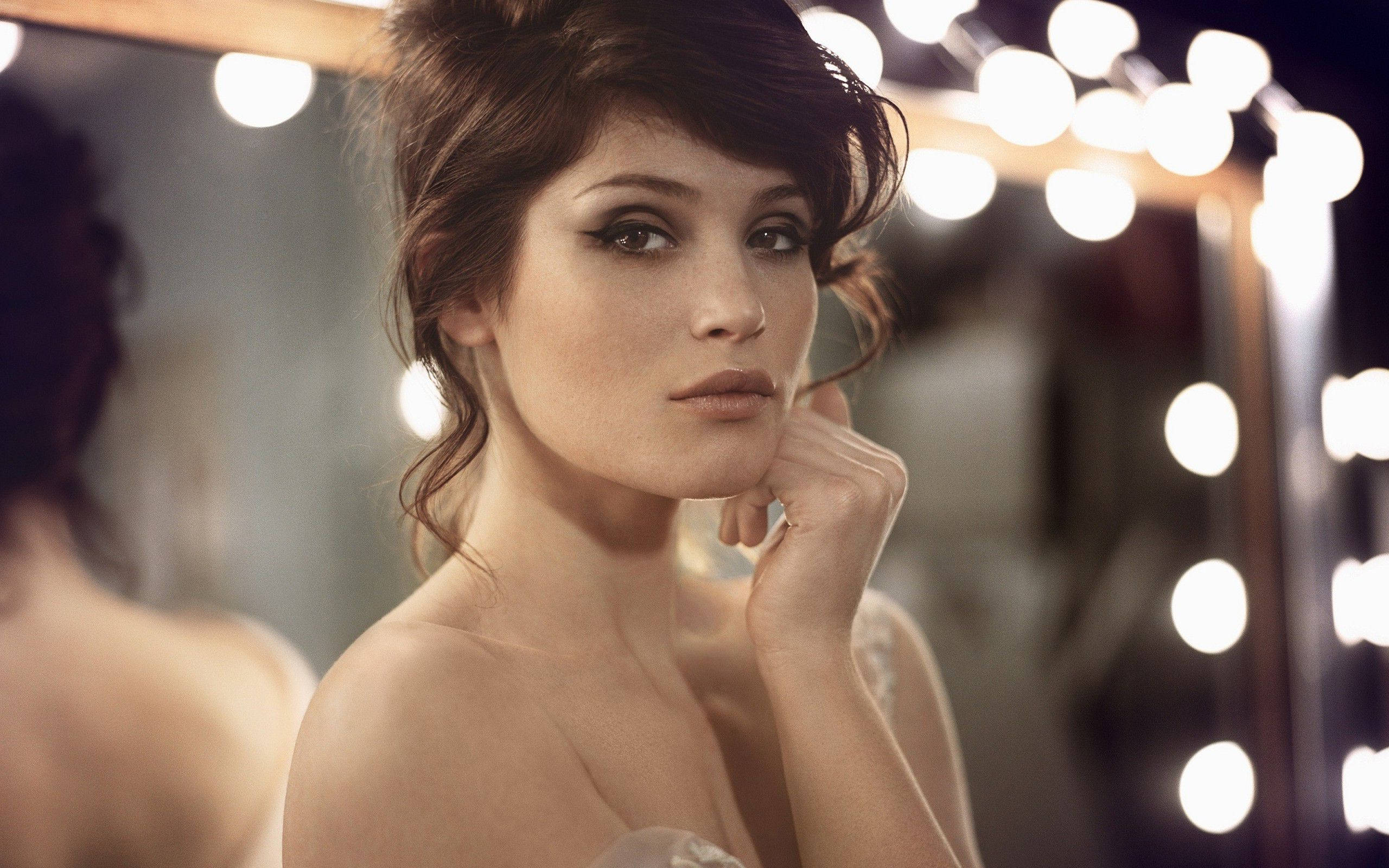 Gemma Arterton 桌布 壁紙 — 一位深色頭髮、煙熏眼妝的女性，在閃爍的化妝燈光下若有所思地凝視著鏡子。