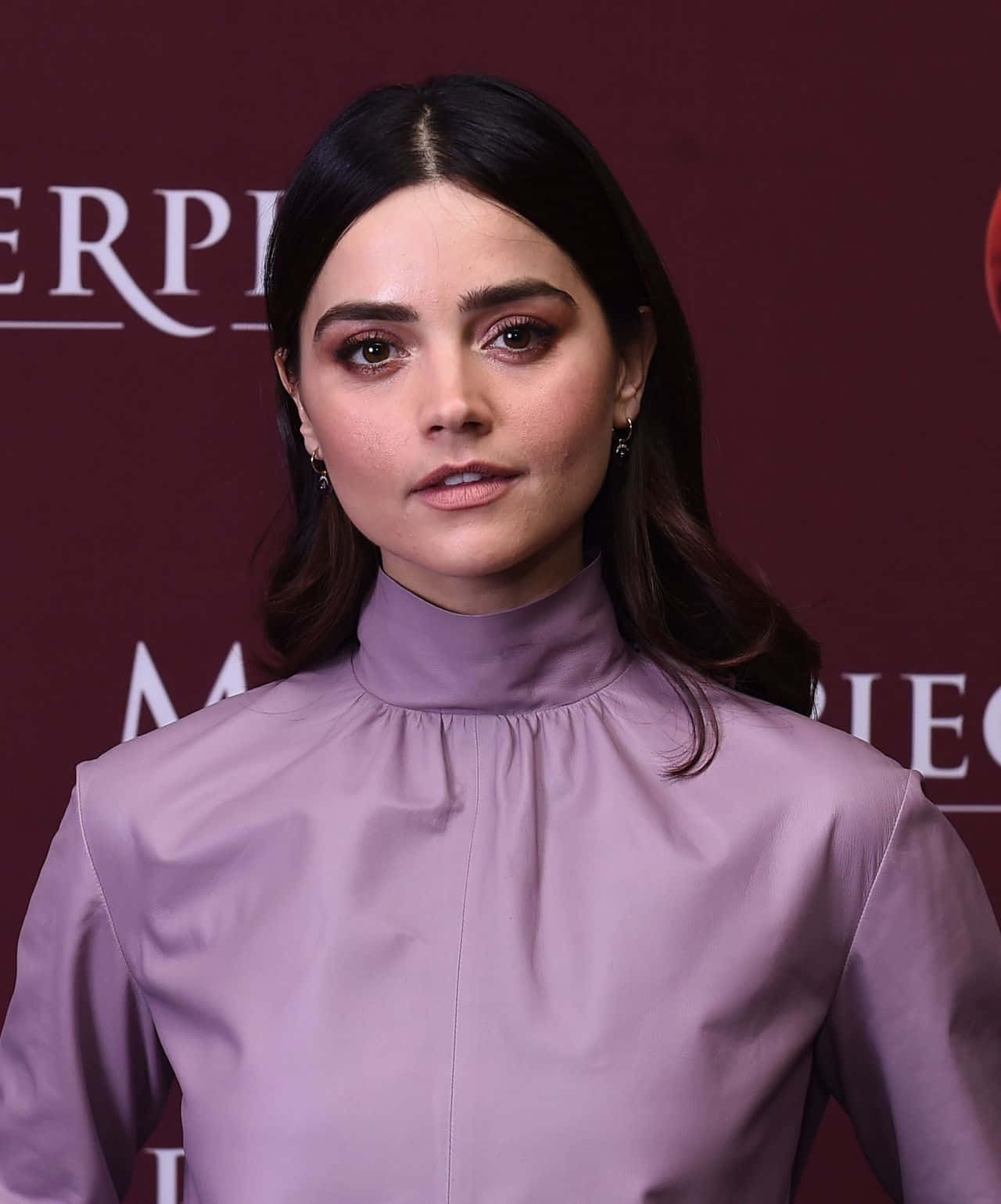 Jenna Coleman 桌布 壁紙 — 一位深色頭髮、煙燻眼妝的女性，在栗色品牌背景前穿著薰衣草色高領襯衫。