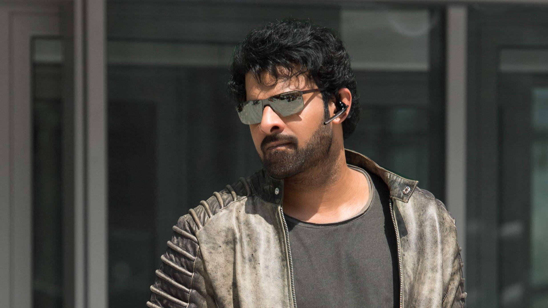 Prabhas HD 桌布 壁紙 — 一位深色頭髮、留鬍鬚的男子戴著太陽眼鏡和一件破舊的皮夾克，站在玻璃門附近的戶外。