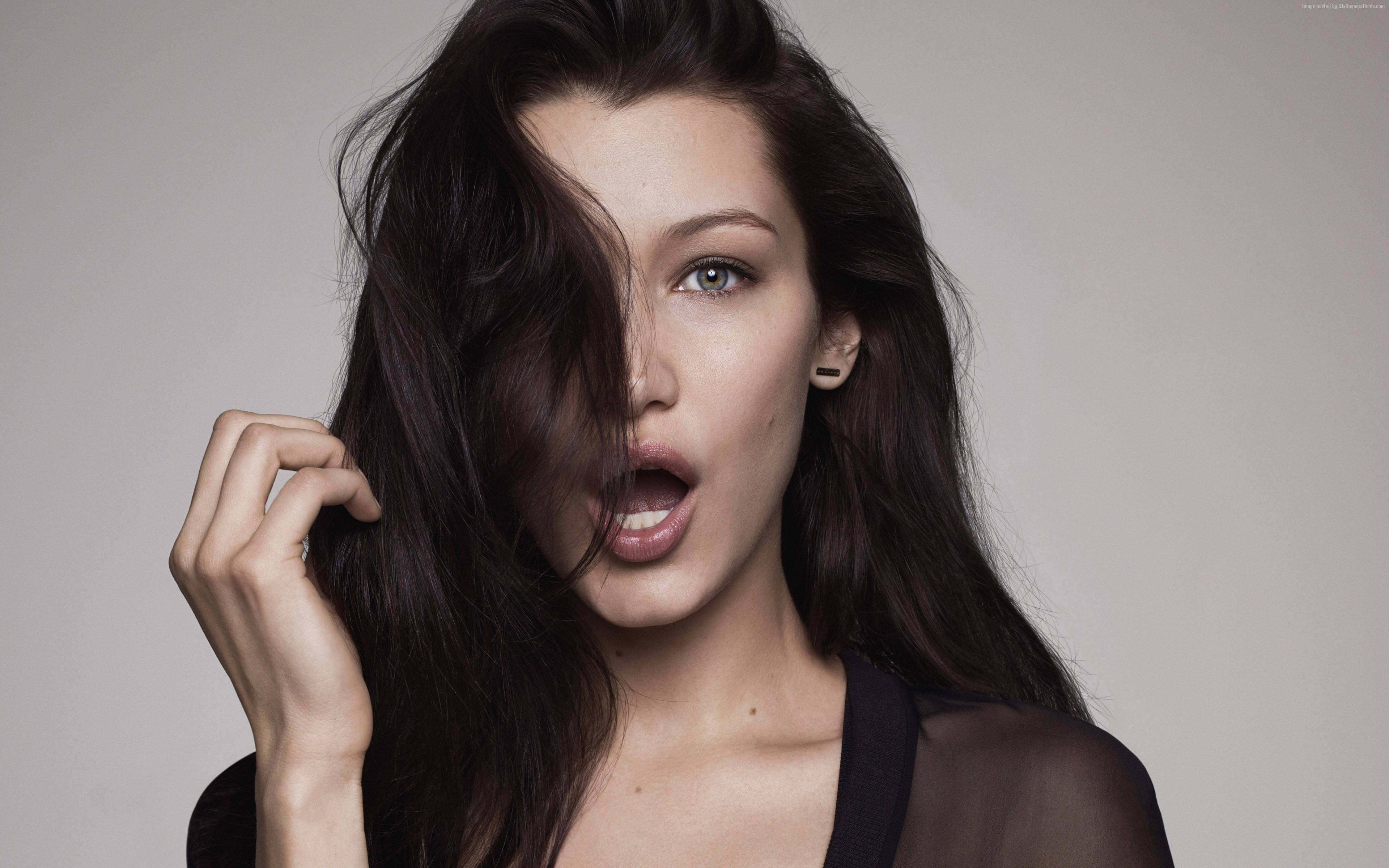 Bella Hadid 桌布 壁紙 — 一位深色頭髮、眼神引人注目的女性，在灰色中性背景下，用手將頭髮從臉上撩起，表情驚訝。
