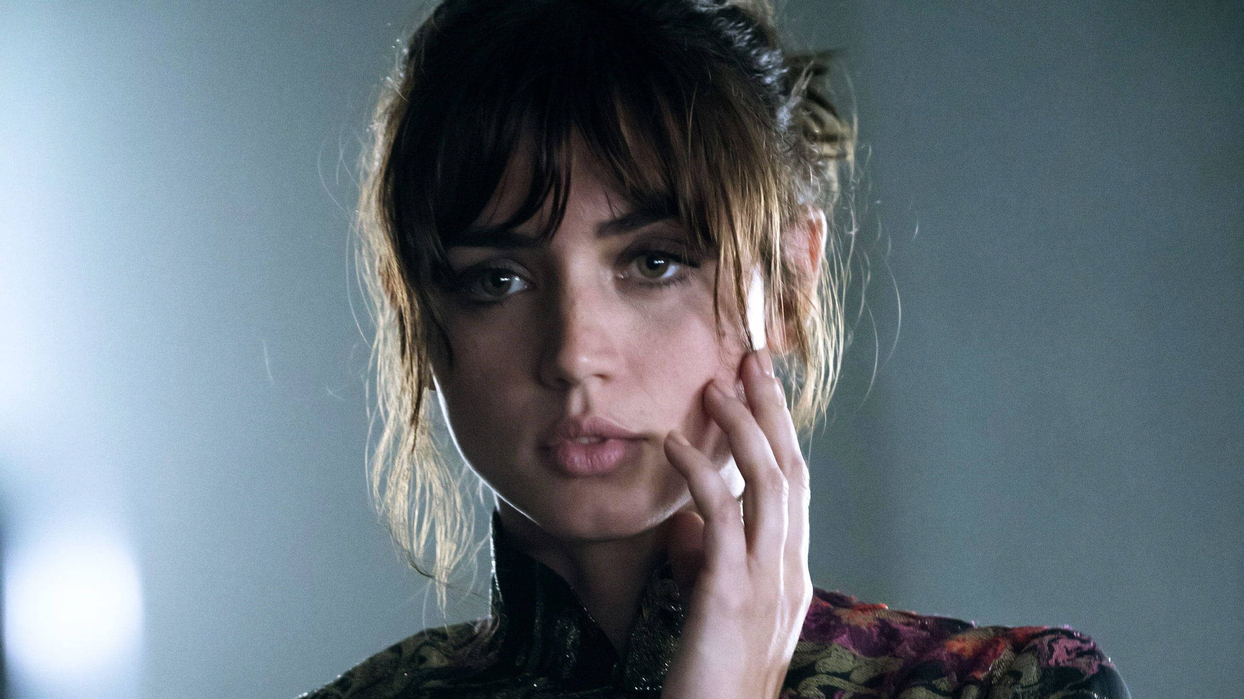 ana de armas 桌布 壁紙 — 一位深色頭髮、眼神強烈的女性，在柔和的灰色背景前觸碰臉頰，直視前方。