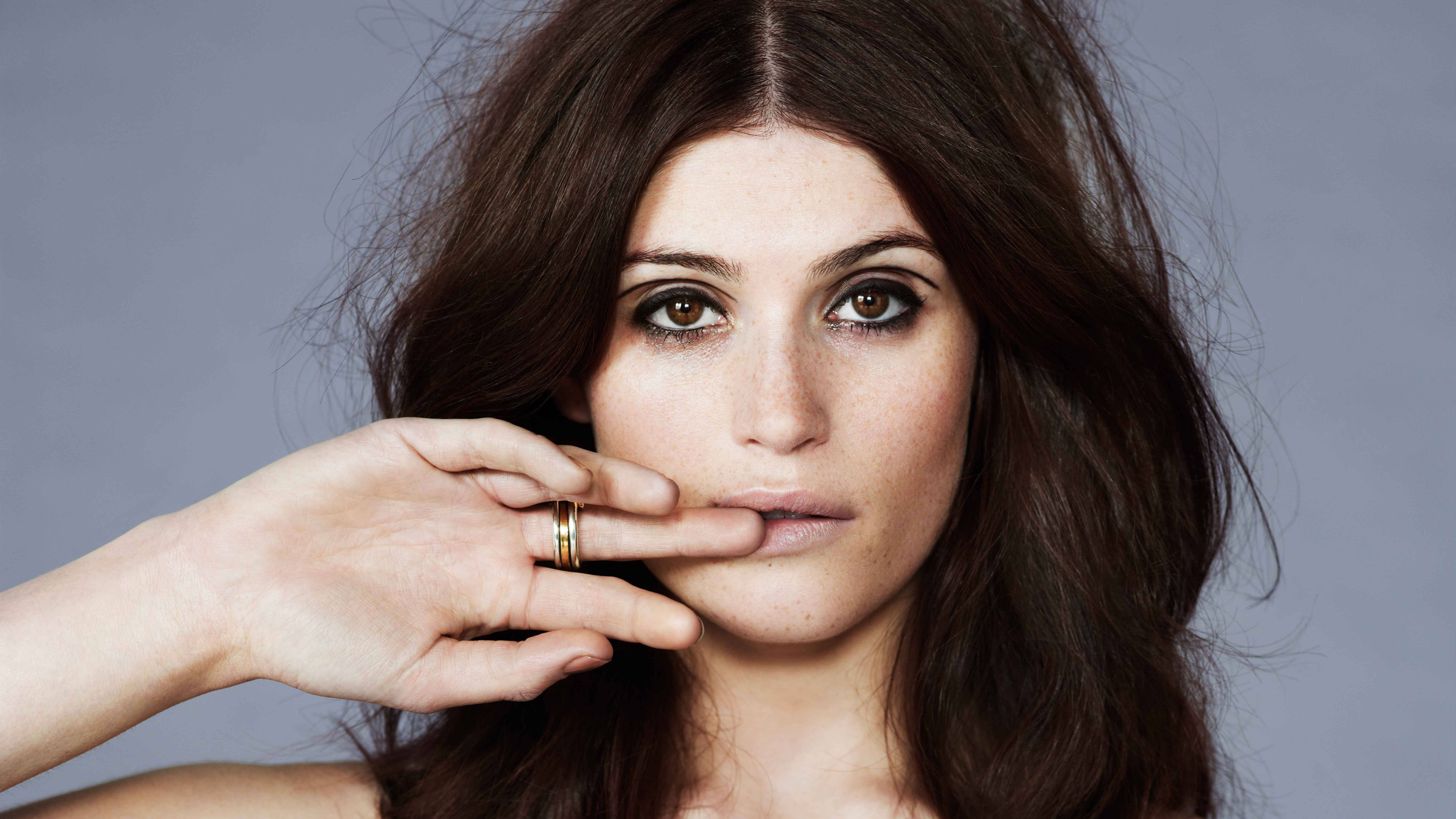 Gemma Arterton 桌布 壁紙 — 一位深色頭髮、眼神強烈的女性，在柔和的灰色背景前，用戴著金戒指的手輕輕遮住嘴唇。