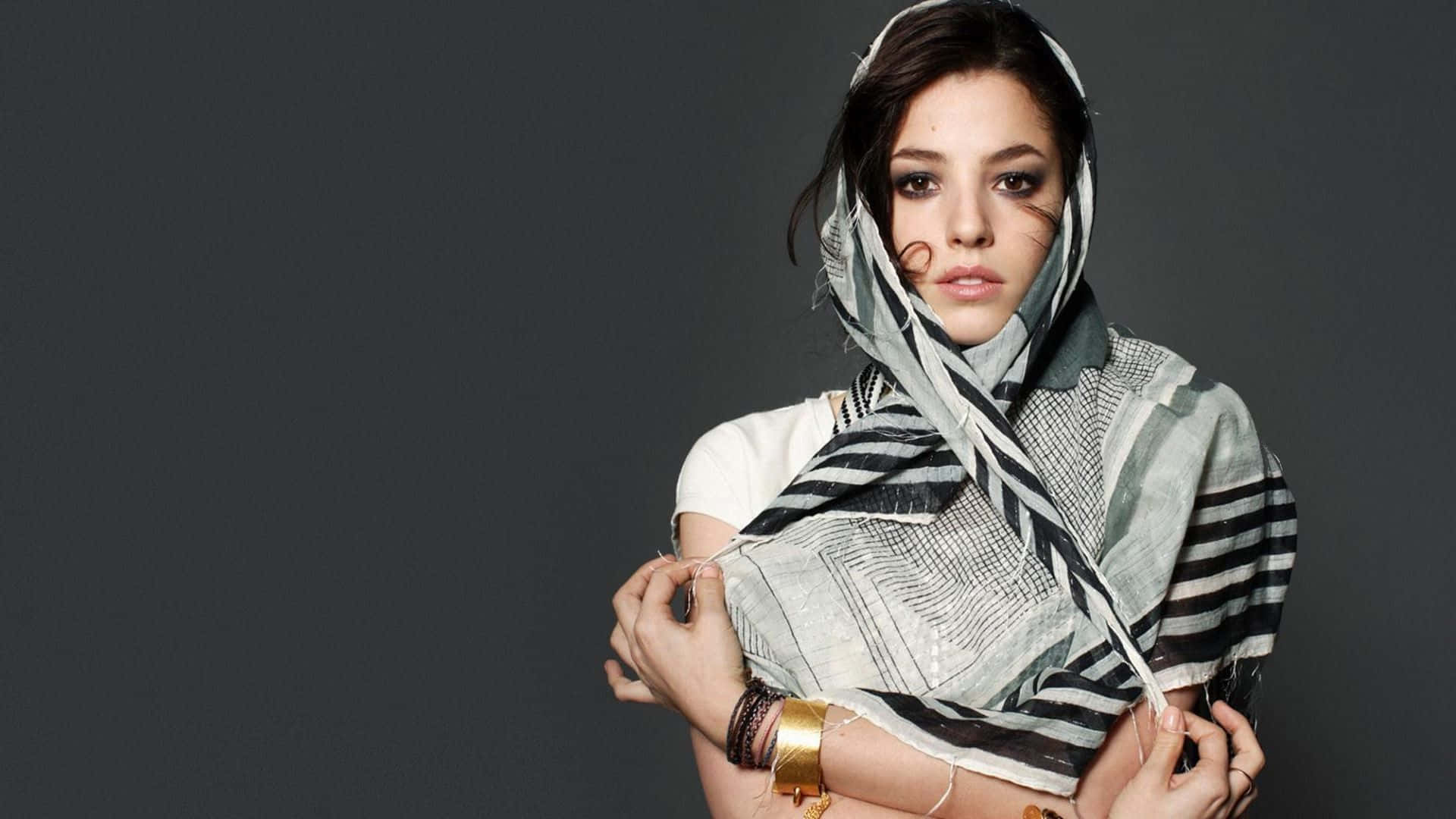 Olivia Thirlby 桌布 壁紙 — 一位深色頭髮、眼神銳利的女性，在純灰色背景前，將一條有圖案的圍巾披在頭上和肩上。