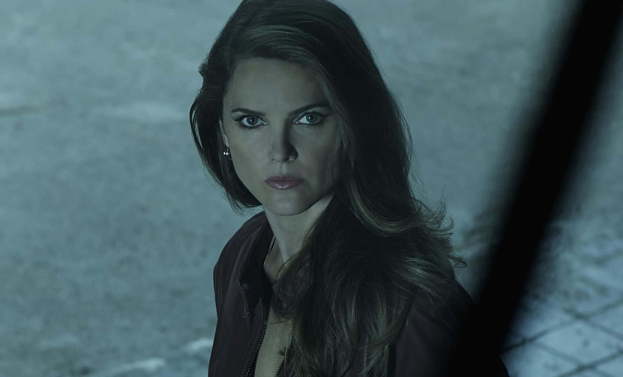 keri russell 桌布 壁紙 — 一位深色頭髮、眼神銳利的女性，在昏暗骯髒的城市小巷中直視前方。
