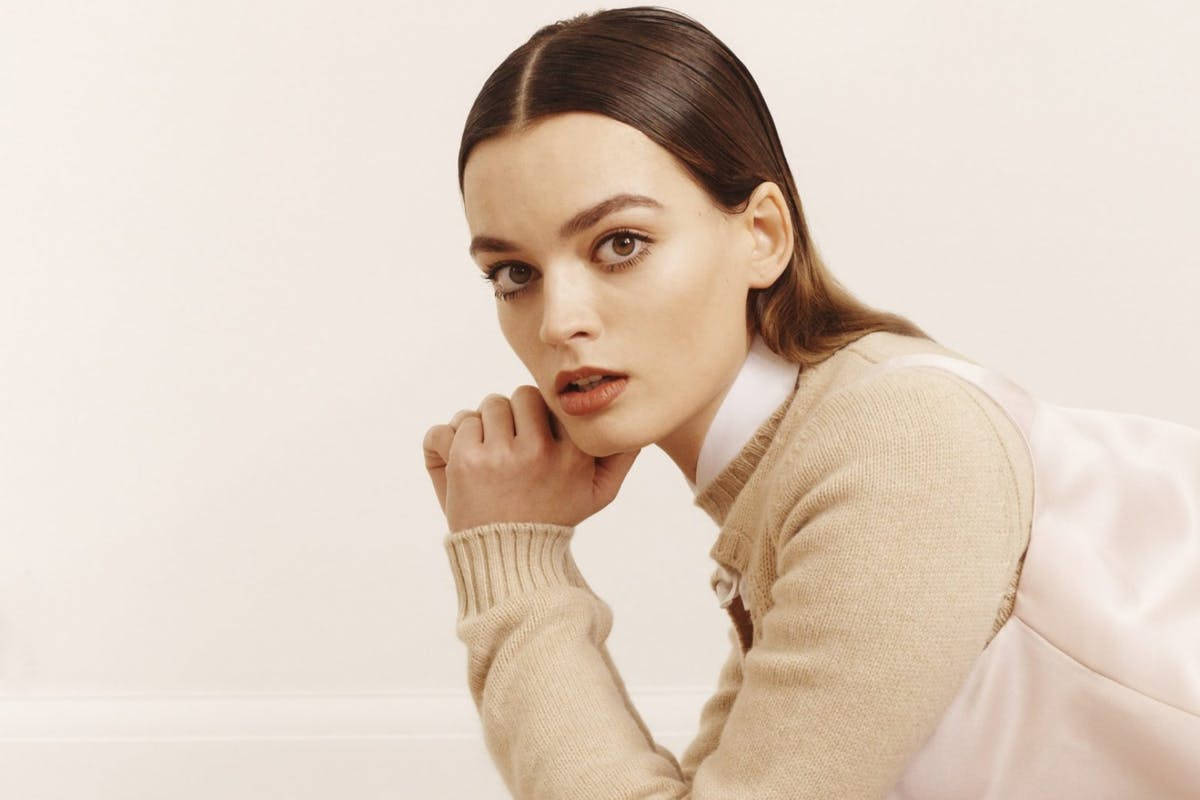maeve wiley 桌布 壁紙 — 一位深色頭髮、眼神銳利的女性，身穿米色毛衣和白色襯衫，在純米白色背景上將下巴擱在手上。