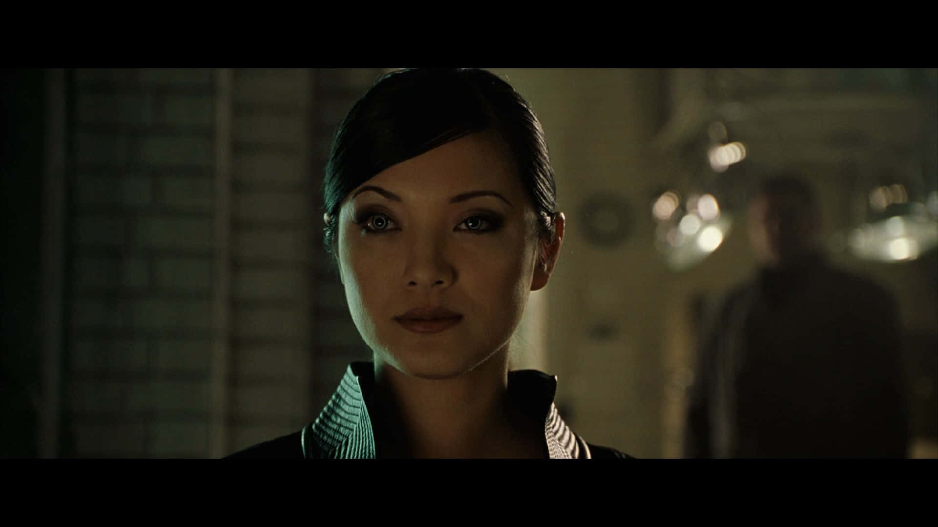 Lady Deathstrike 桌布 壁紙 — 一位深色頭髮、眼神銳利的女性在昏暗的房間中向前凝視，背景有模糊的燈光和一個人影。
