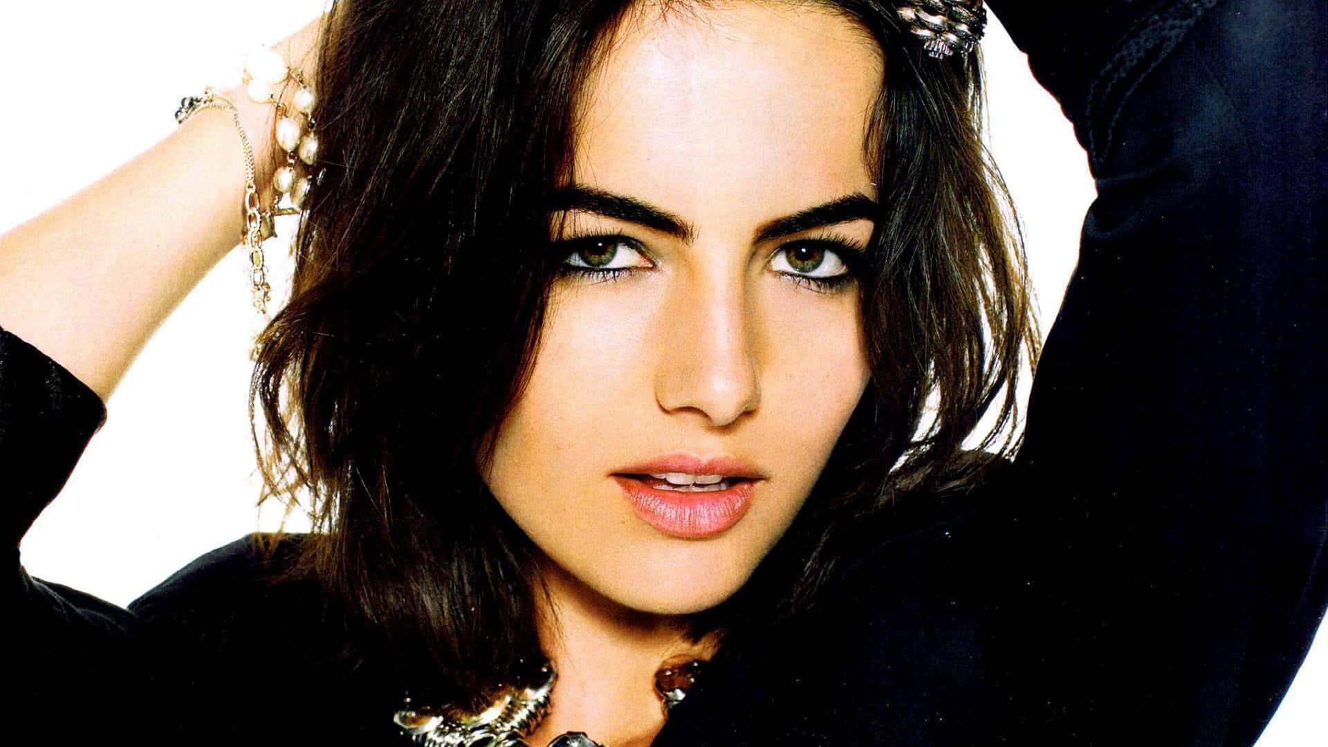 Camilla Belle 桌布 壁紙 — 一位深色頭髮、眼神銳利的女性特寫肖像，佩戴珠寶，身穿黑色上衣，背景為白色。