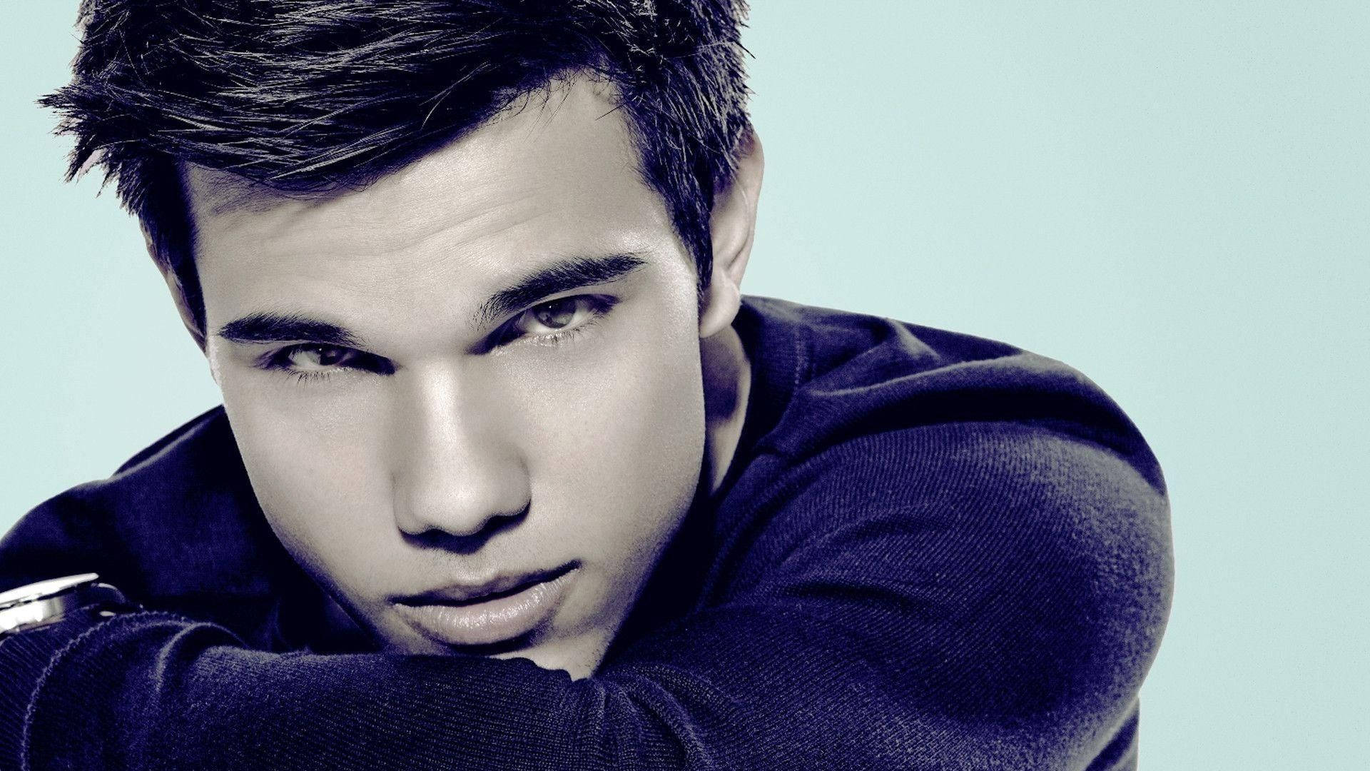 Taylor Lautner 桌布 壁紙 — 一位深色頭髮、眼神銳利的年輕男子，身穿深色毛衣，在淺藍色背景前向前傾身。