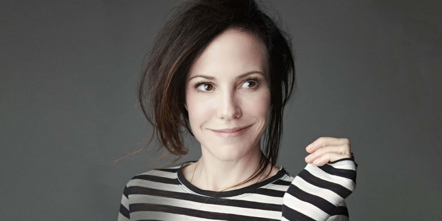 mary louise parker 桌布 壁紙 — 一位深色頭髮、穿著條紋襯衫的女性，在灰色背景下微笑著望向遠方。