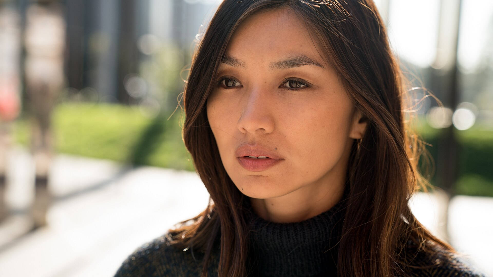Gemma Chan 桌布 壁紙 — 一位深色頭髮、穿著黑色高領毛衣的女性在晴朗的日子裡若有所思地望著戶外，身後是模糊的城市綠化。