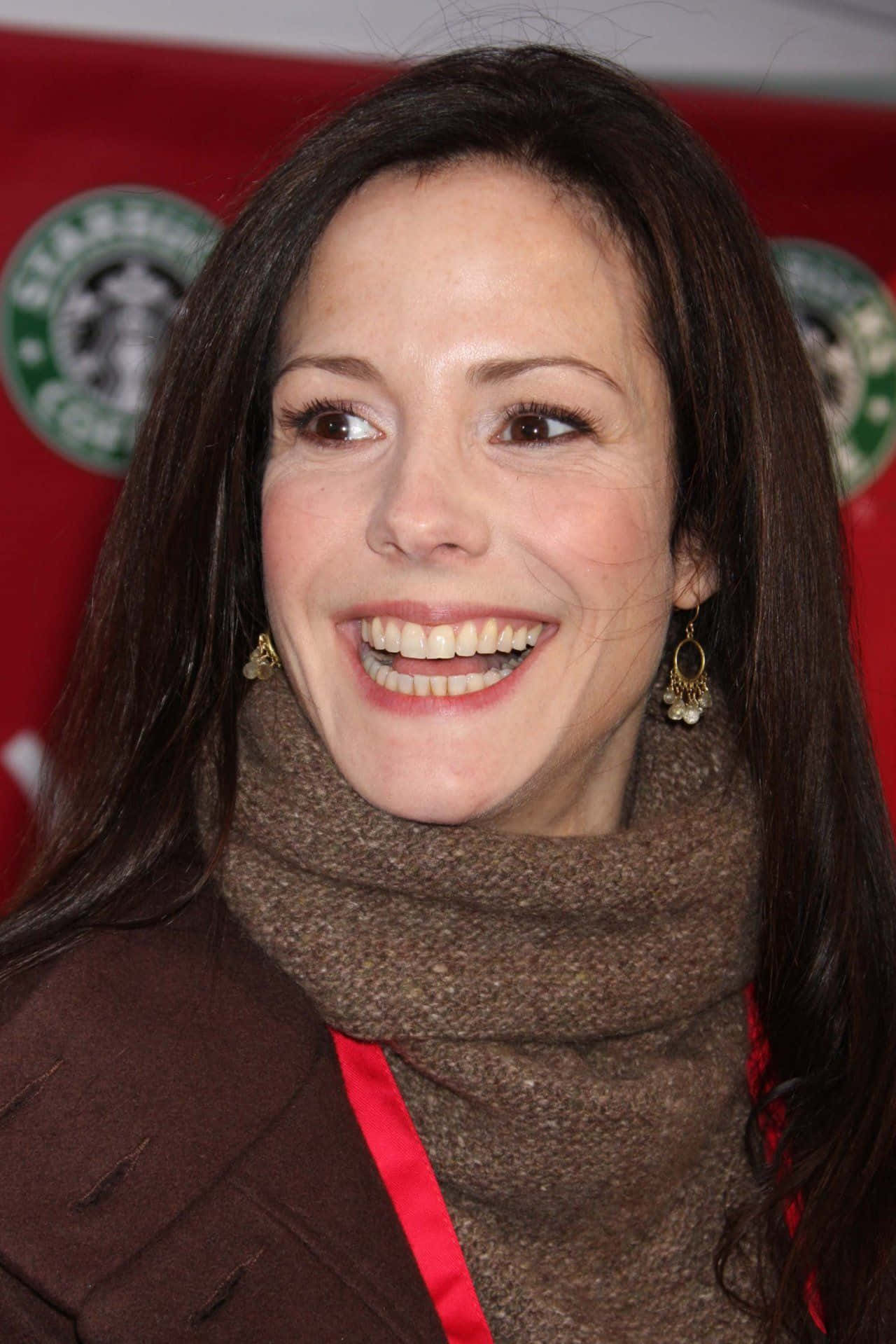 mary louise parker 桌布 壁紙 — 一位深色頭髮、笑容溫暖的女性，在紅色Starbucks背景下戴著棕色圍巾和金色耳環。