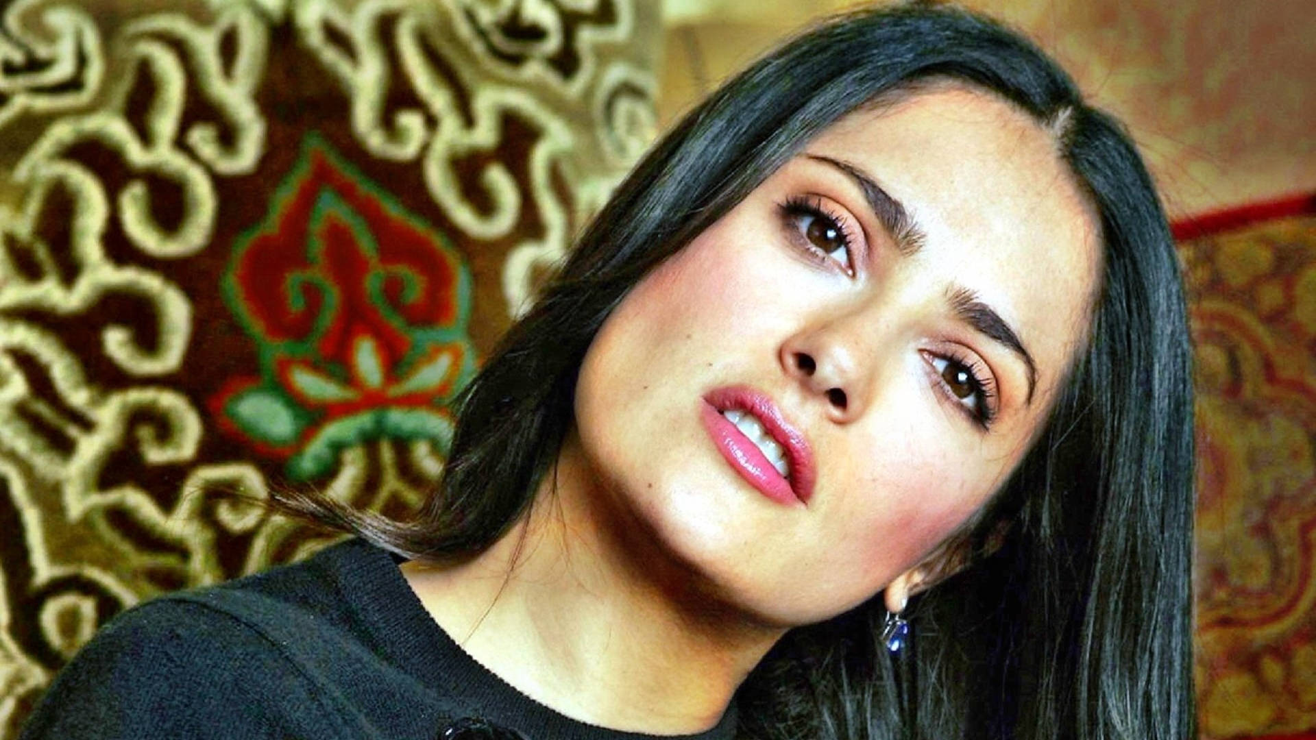 Salma 桌布 壁紙 — 一位深色頭髮、粉紅嘴唇的女性，在華麗圖案掛毯背景下向上凝視。