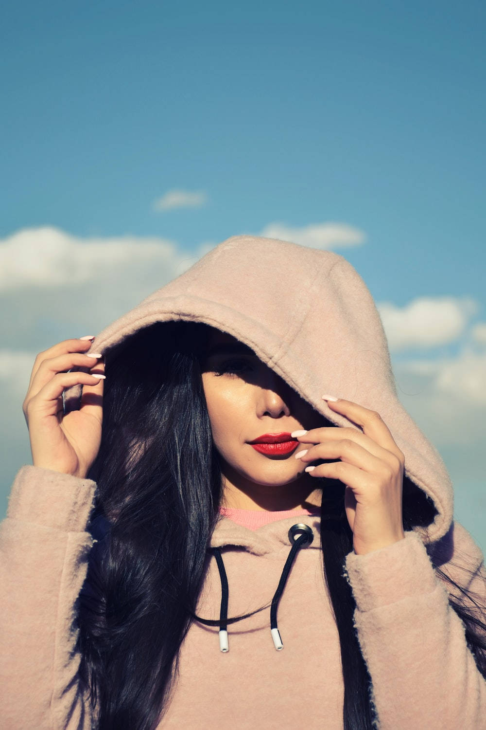 Hoodie 桌布 壁紙 — 一位深色頭髮、紅唇的女性在藍天白雲下調整她的粉紅色連帽衫。