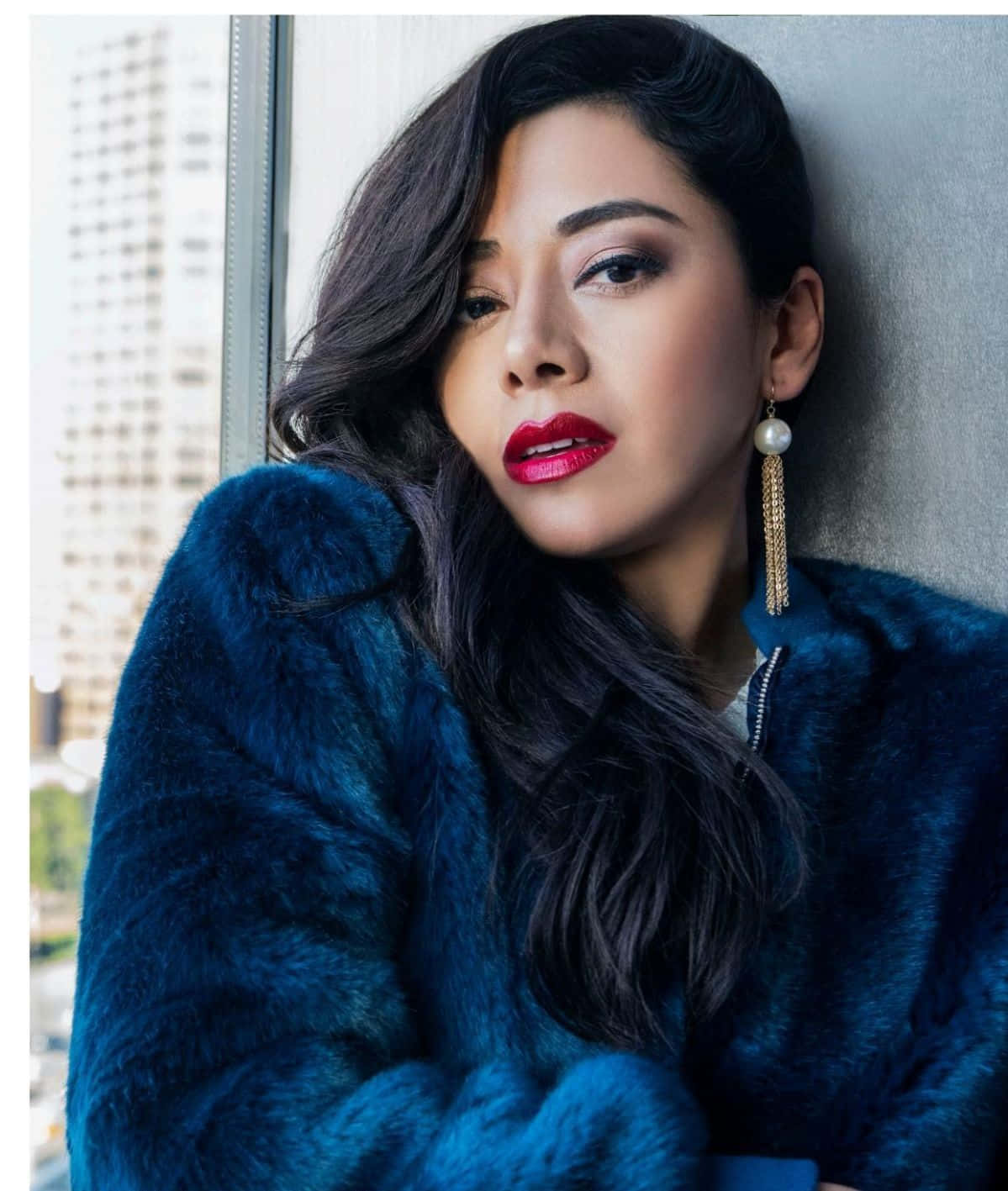 Aimee Garcia 桌布 壁紙 — 一位深色頭髮、紅唇的女性穿著藍色毛皮大衣，靠著窗戶，身後可見城市建築。