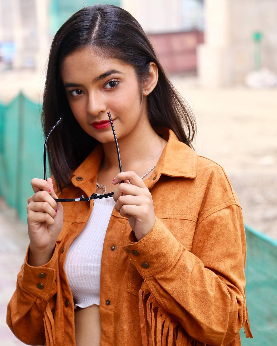 Indian 桌布 壁紙 — 一位深色頭髮、紅唇的年輕女性，身穿橙色流蘇外套，手持黑色眼鏡，在戶外。