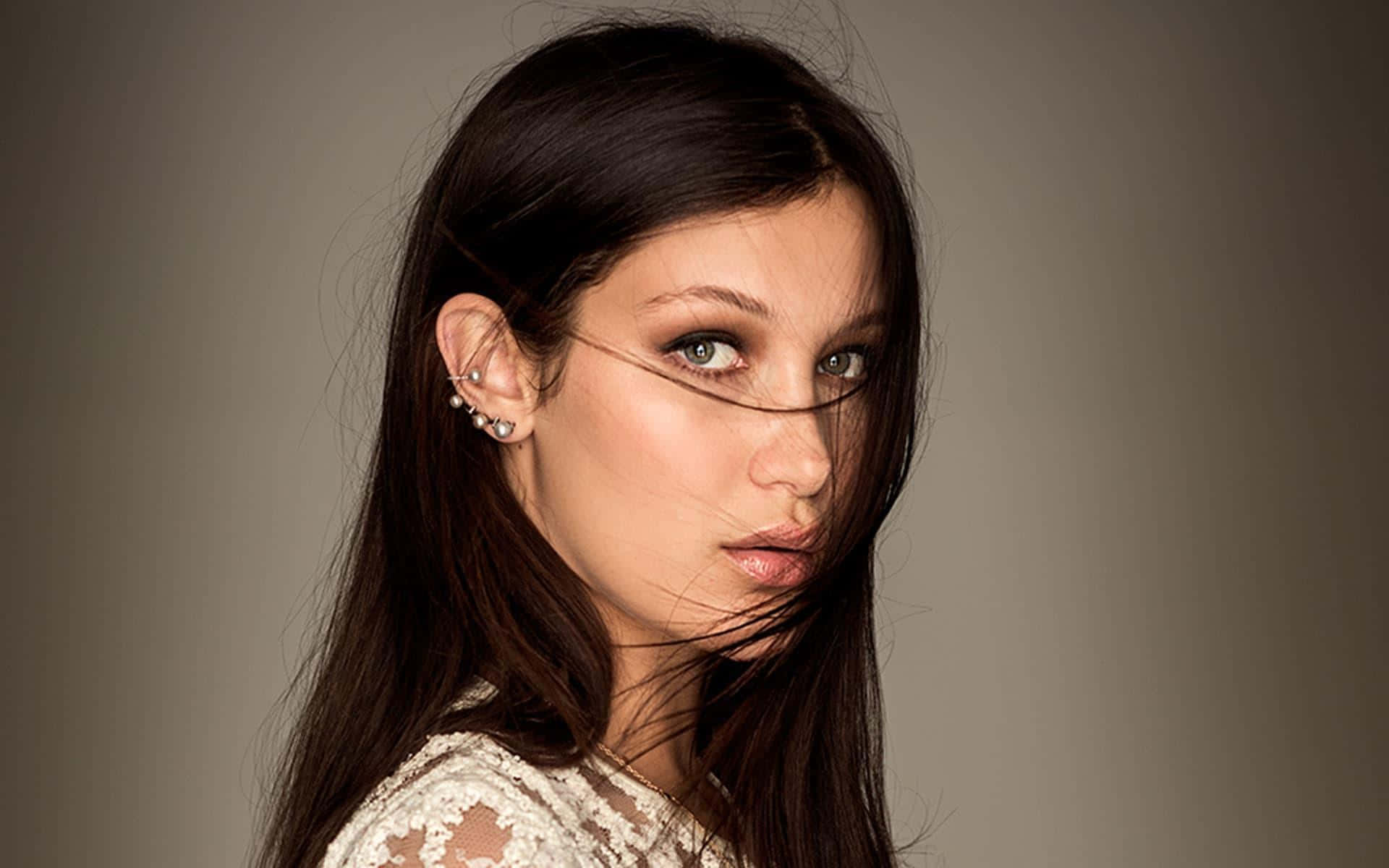 Bella Hadid 桌布 壁紙 — 一位深色頭髮、綠色眼睛的女性在灰色背景下，風吹亂她的髮絲，她凝視著鏡頭，眼神強烈。