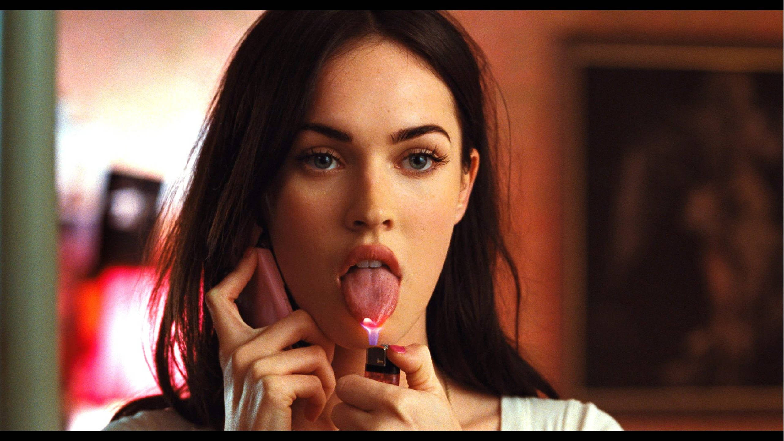Megan Fox 桌布 壁紙 — 一位深色頭髮、藍色眼睛的女性，將電話貼在耳邊，同時舔著點燃的打火機火焰。