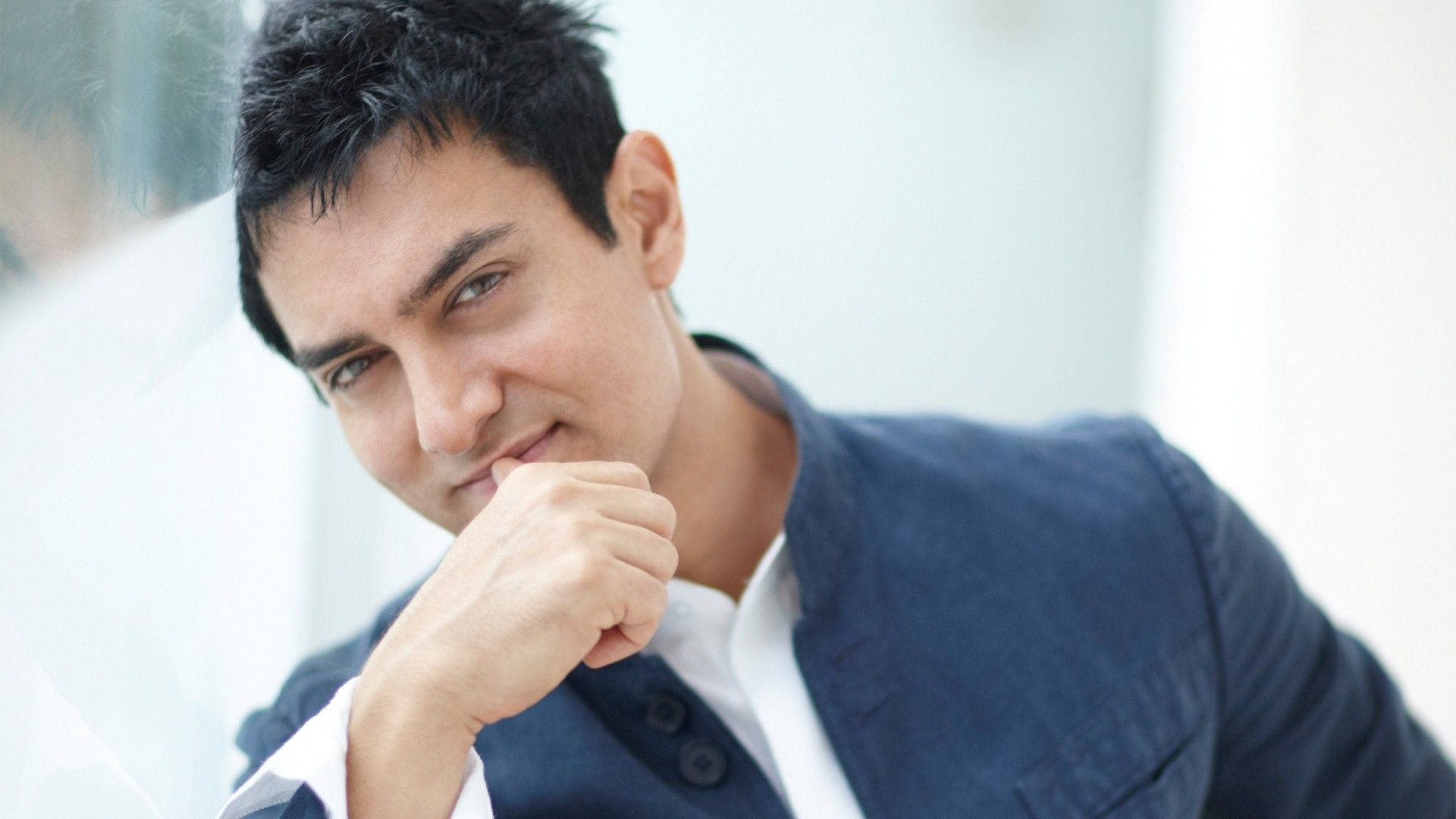 Aamir Khan 桌布 壁紙 — 一位深色頭髮、藍色眼睛的男子，在明亮的白色背景前，將下巴擱在拳頭上，溫柔地微笑。