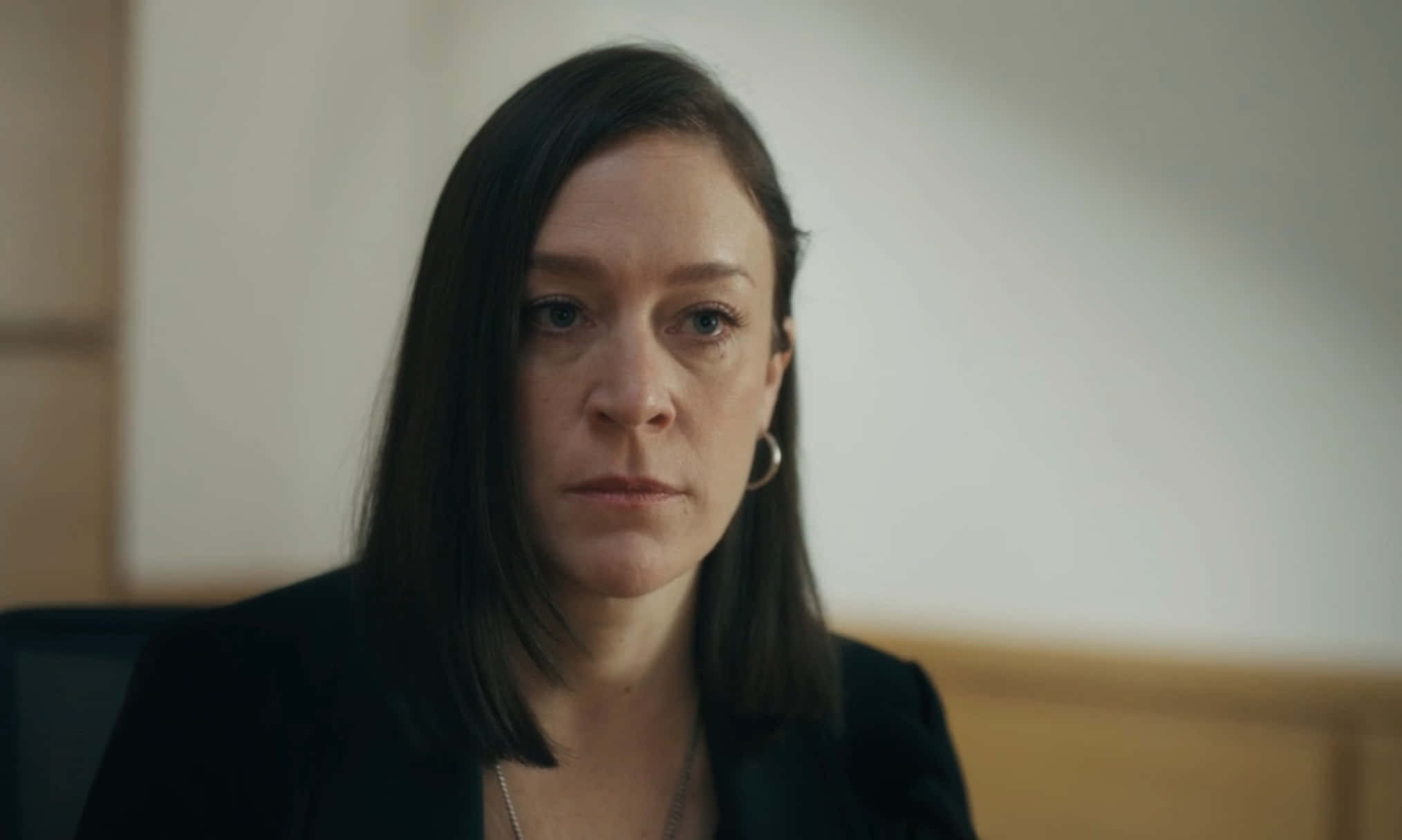 Chloë Sevigny 桌布 壁紙 — 一位深色頭髮、表情嚴肅的女性，穿著黑色西裝外套，坐在柔和光線的室內環境中。