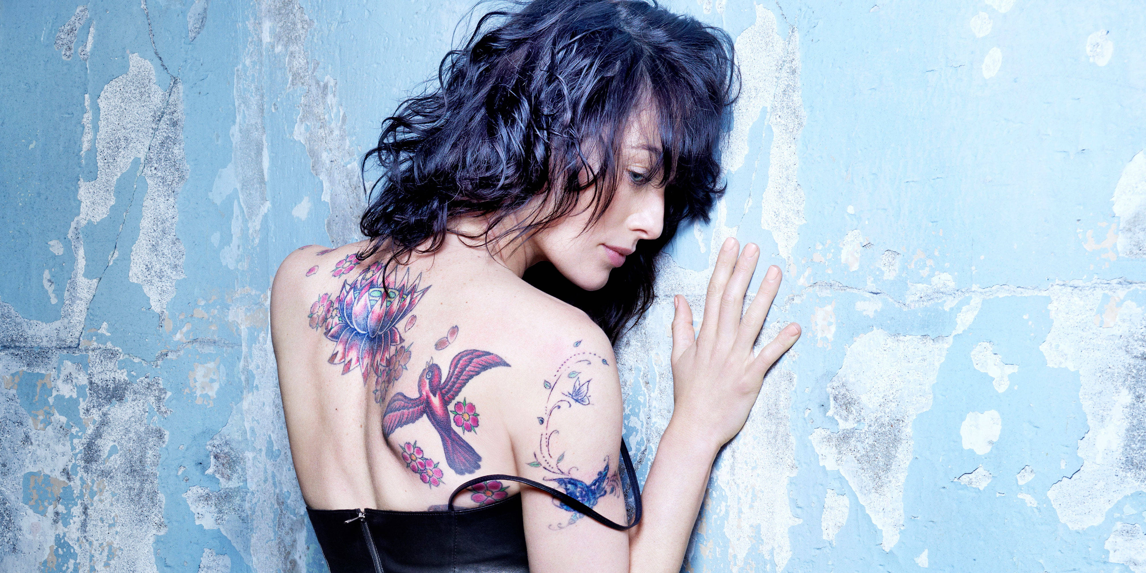 Tattoo 桌布 壁紙 — 一位深色頭髮、身上有彩色刺青的女子靠著一面風化的藍色牆壁，手放在牆面上。