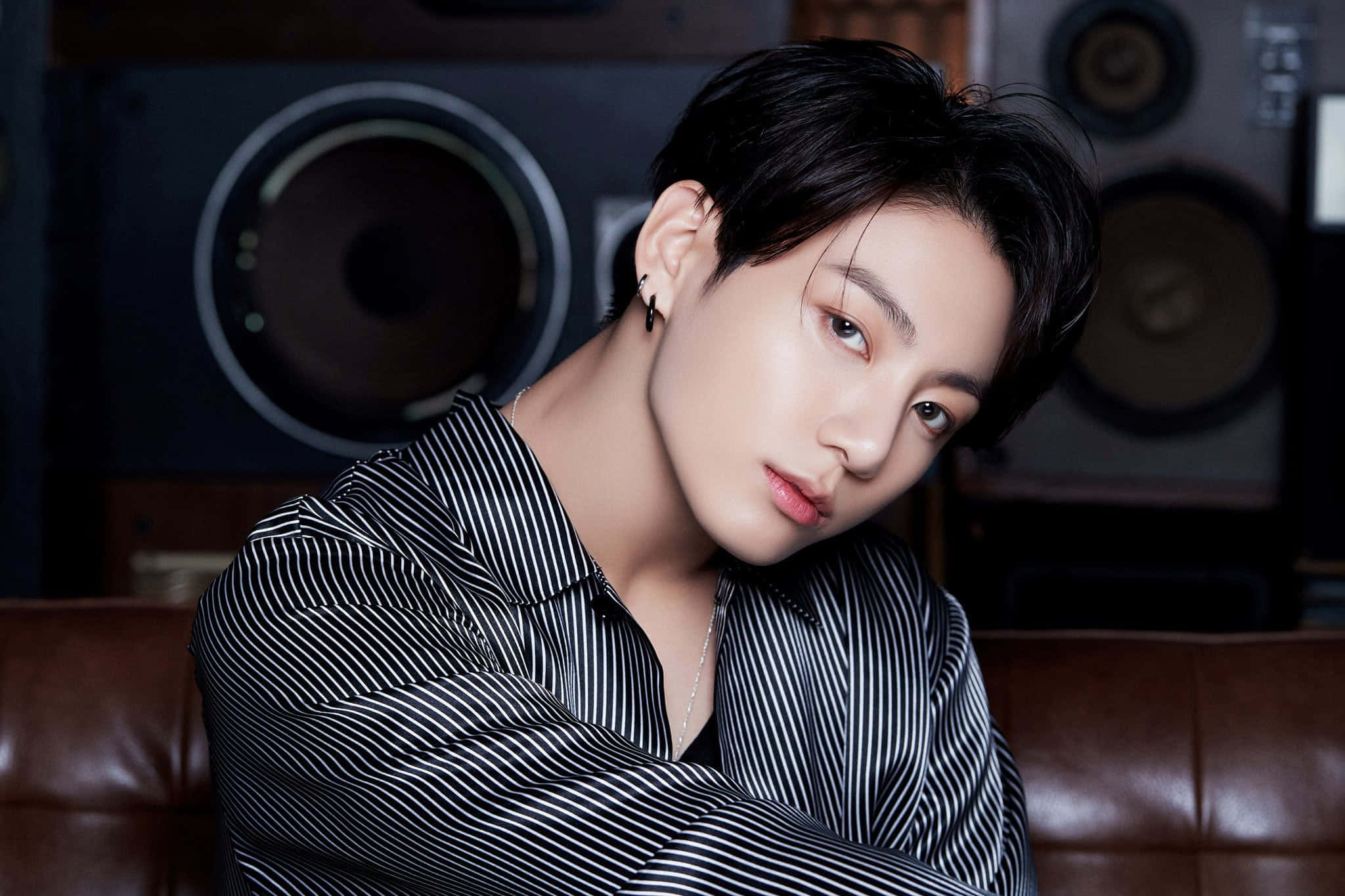Jungkook 桌布 壁紙 — 一位深色頭髮、身穿條紋襯衫的年輕男子，在復古喇叭的背景襯托下，自信地坐在棕色皮沙發上。
