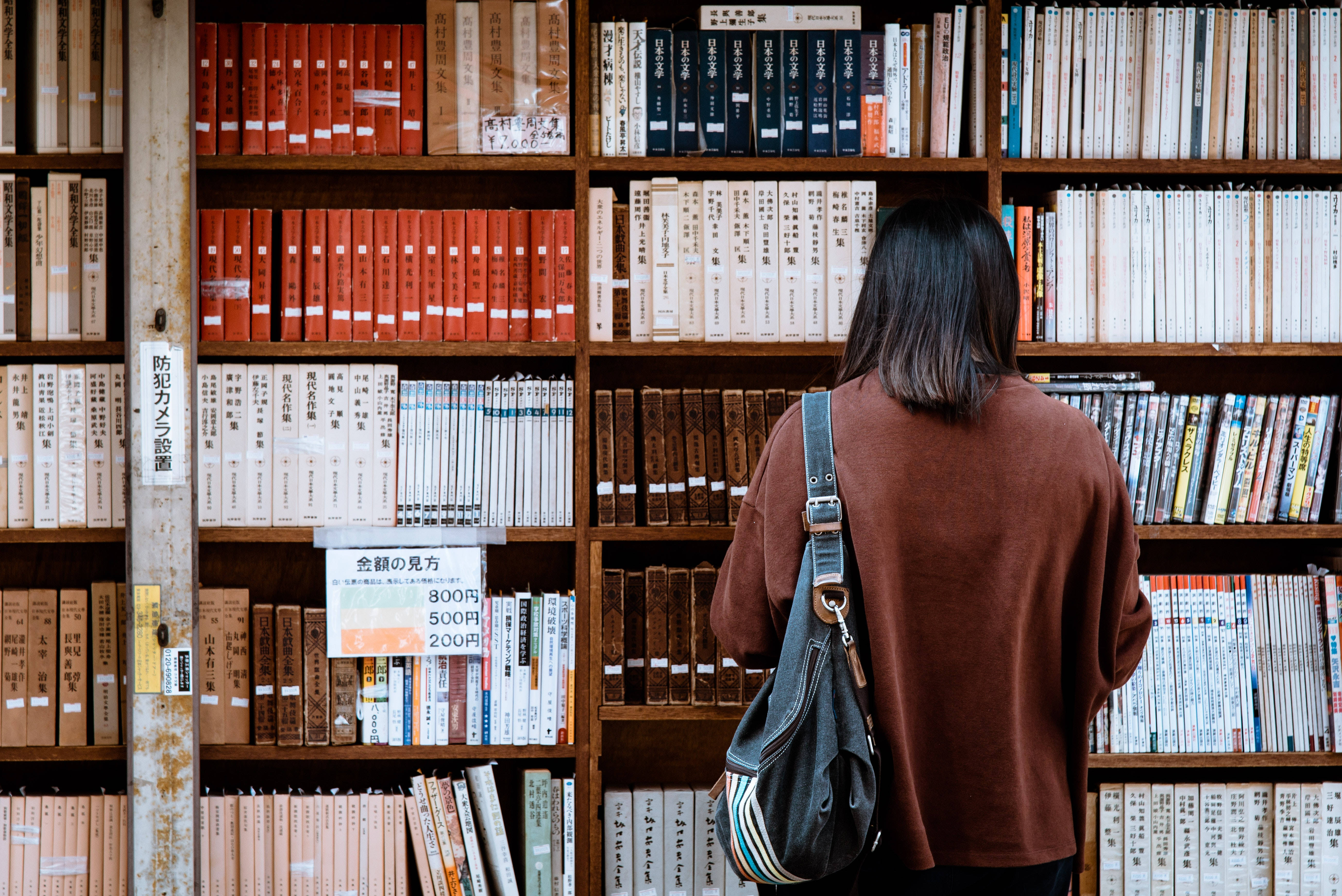 讀書動力 桌布 壁紙 — 一位深色頭髮、身穿棕色毛衣的人正在木製書店通道中瀏覽書籍，書架上擺滿了色彩繽紛的書脊。