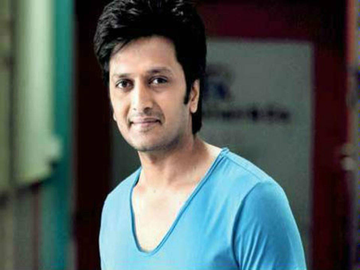 riteish deshmukh 桌布 壁紙 — 一位深色頭髮、身穿淺藍色V領襯衫的男子，在柔和模糊的室內環境中對著鏡頭溫柔微笑。