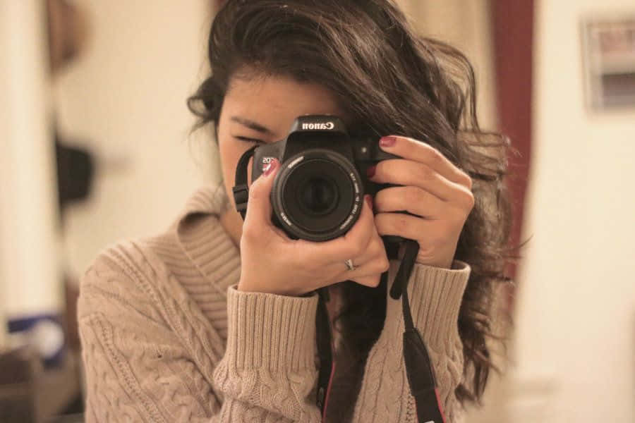 Holding Camera 桌布 壁紙 — 一位深色頭髮、身穿米色毛衣的女性，在溫暖的室內環境中將黑色 Canon DSLR 相機舉到臉前。