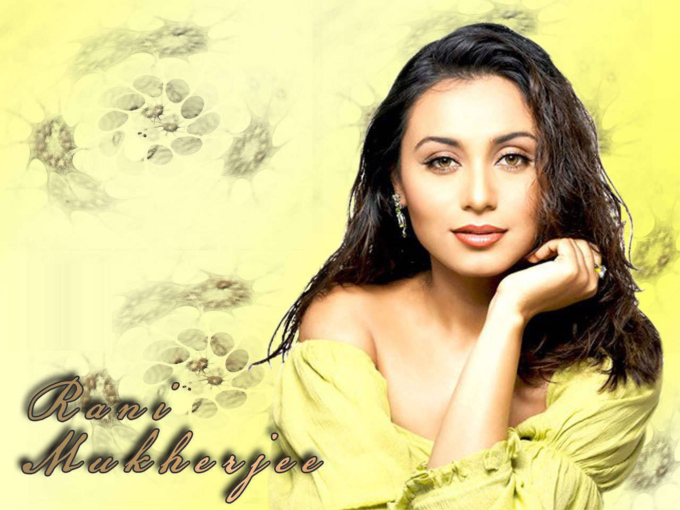 Rani Mukerji 桌布 壁紙 — 一位深色頭髮、身穿綠色上衣的女性，在柔和的黃色花卉背景前優雅地擺姿勢。