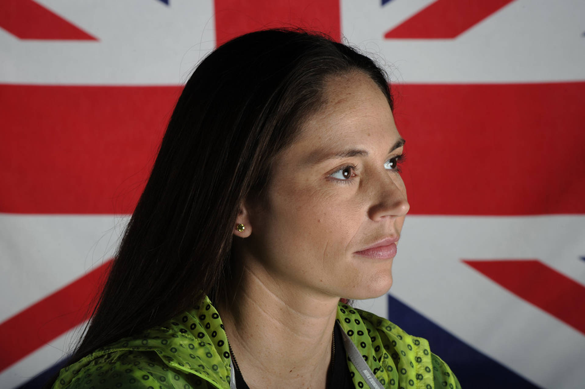 Sue 桌布 壁紙 — 一位深色頭髮、身穿綠色圖案外套的女性，在模糊的英國國旗背景下若有所思地凝視。