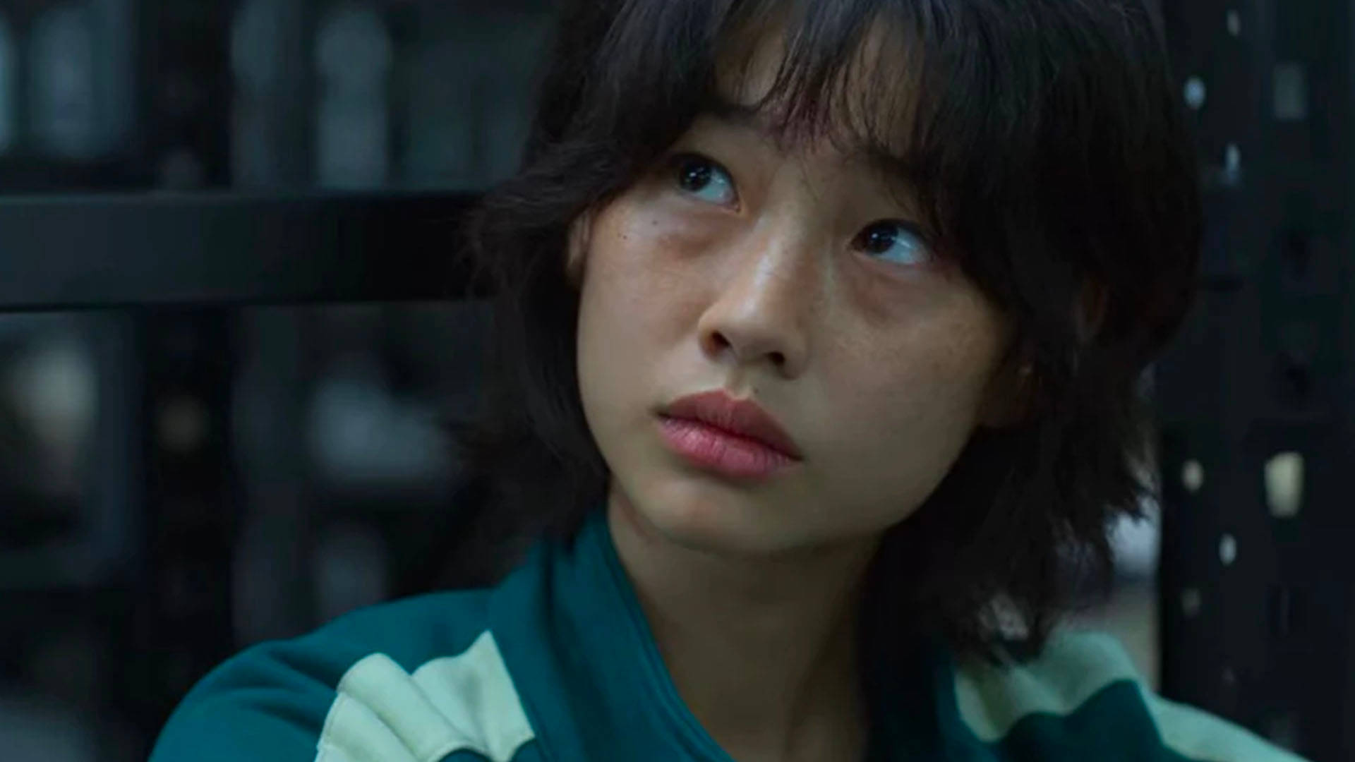 桌布 壁紙 — 一位深色頭髮、身穿綠色外套的年輕女性在昏暗的室內環境中抬頭向上看，表情嚴肅。