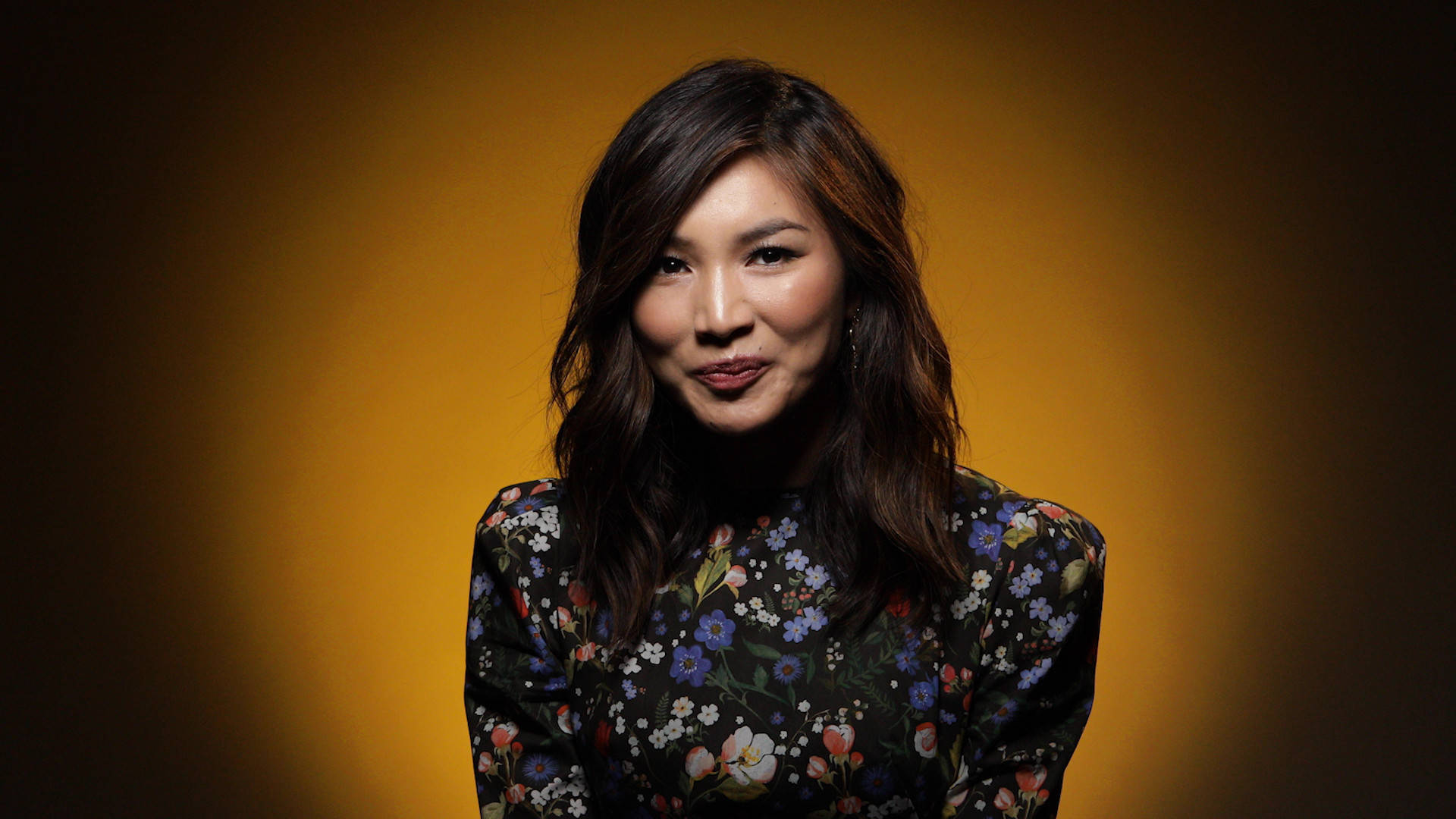 Gemma Chan 桌布 壁紙 — 一位深色頭髮、身穿花卉圖案襯衫的女性，在溫暖的金色漸變背景前溫柔微笑。
