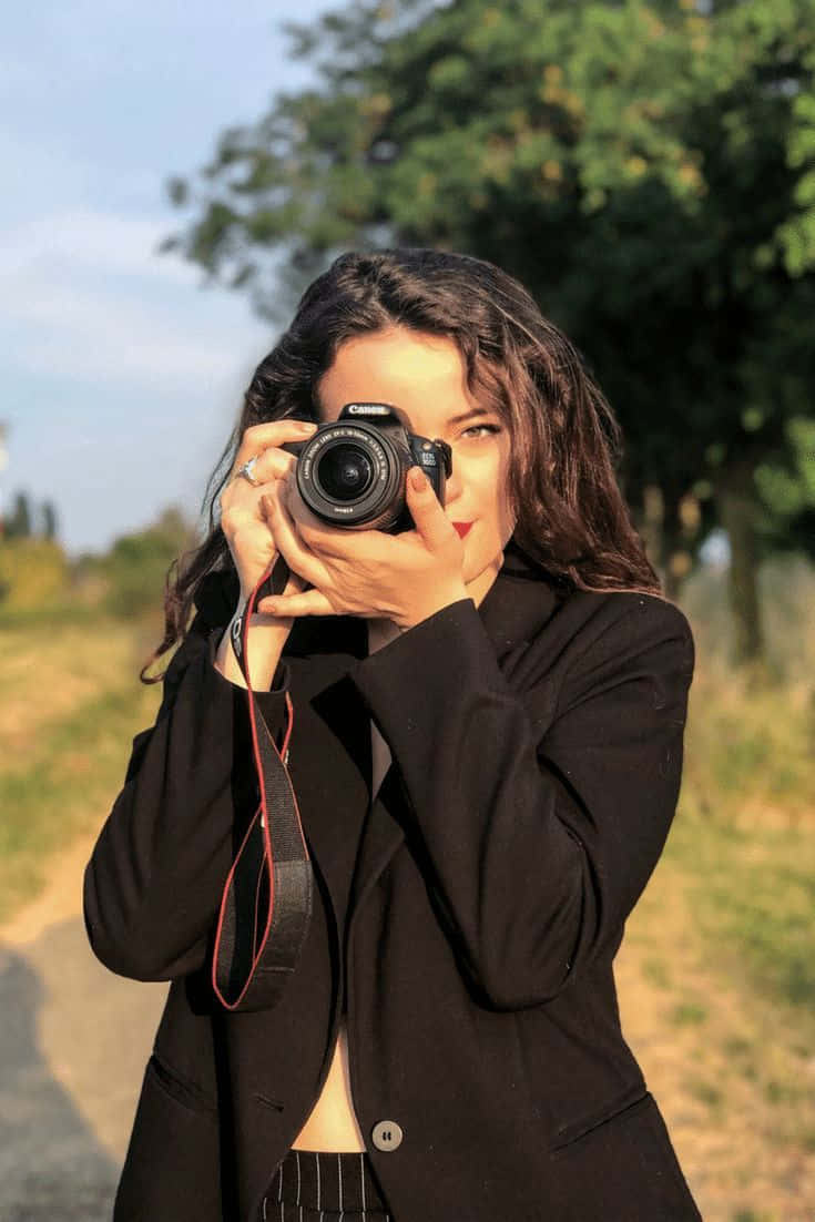 Holding Camera 桌布 壁紙 — 一位深色頭髮、身穿黑色西裝外套的女性，在明亮的陽光下於戶外將單反相機舉到臉前。