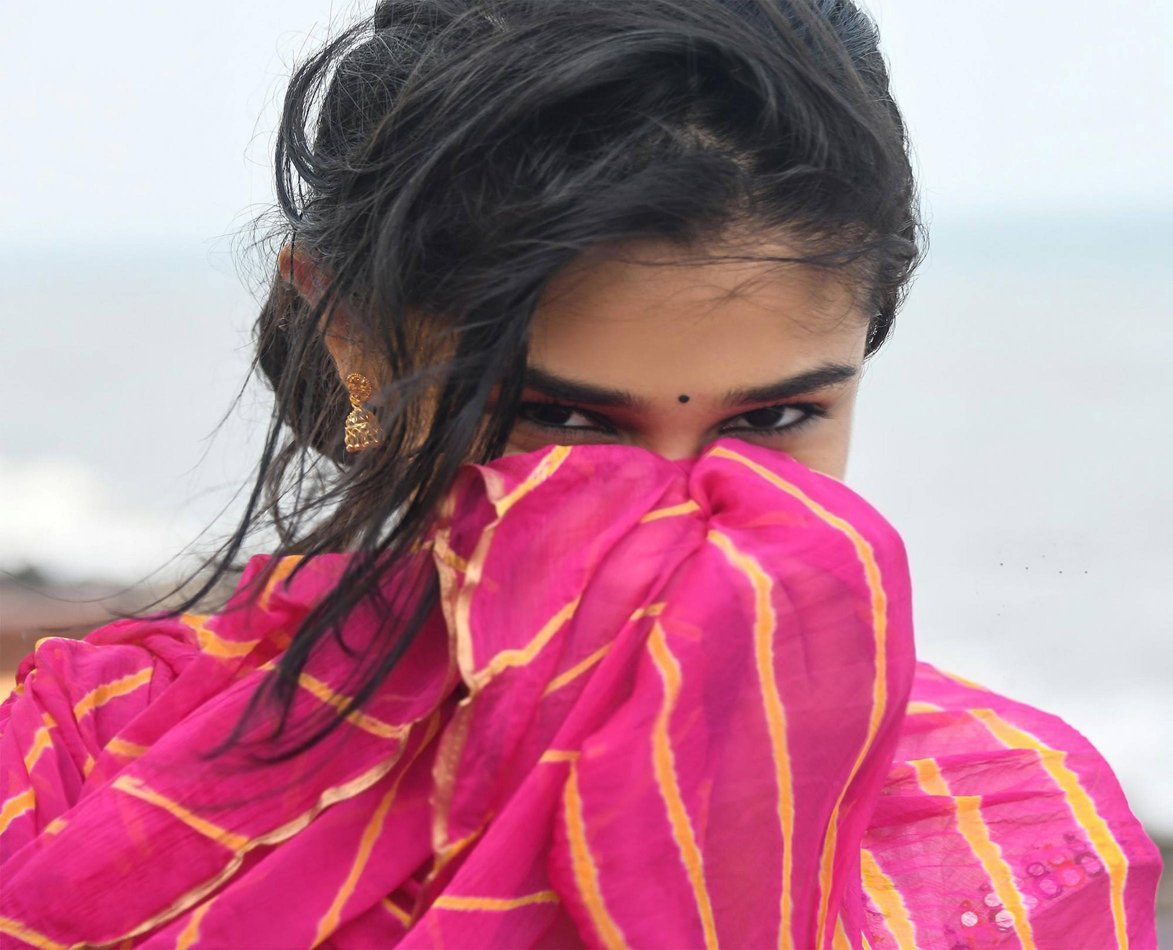 Indian 桌布 壁紙 — 一位深色頭髮、額頭有紅點的年輕女性，在模糊的海岸背景下，透過一條鮮豔的粉黃條紋圍巾窺視。