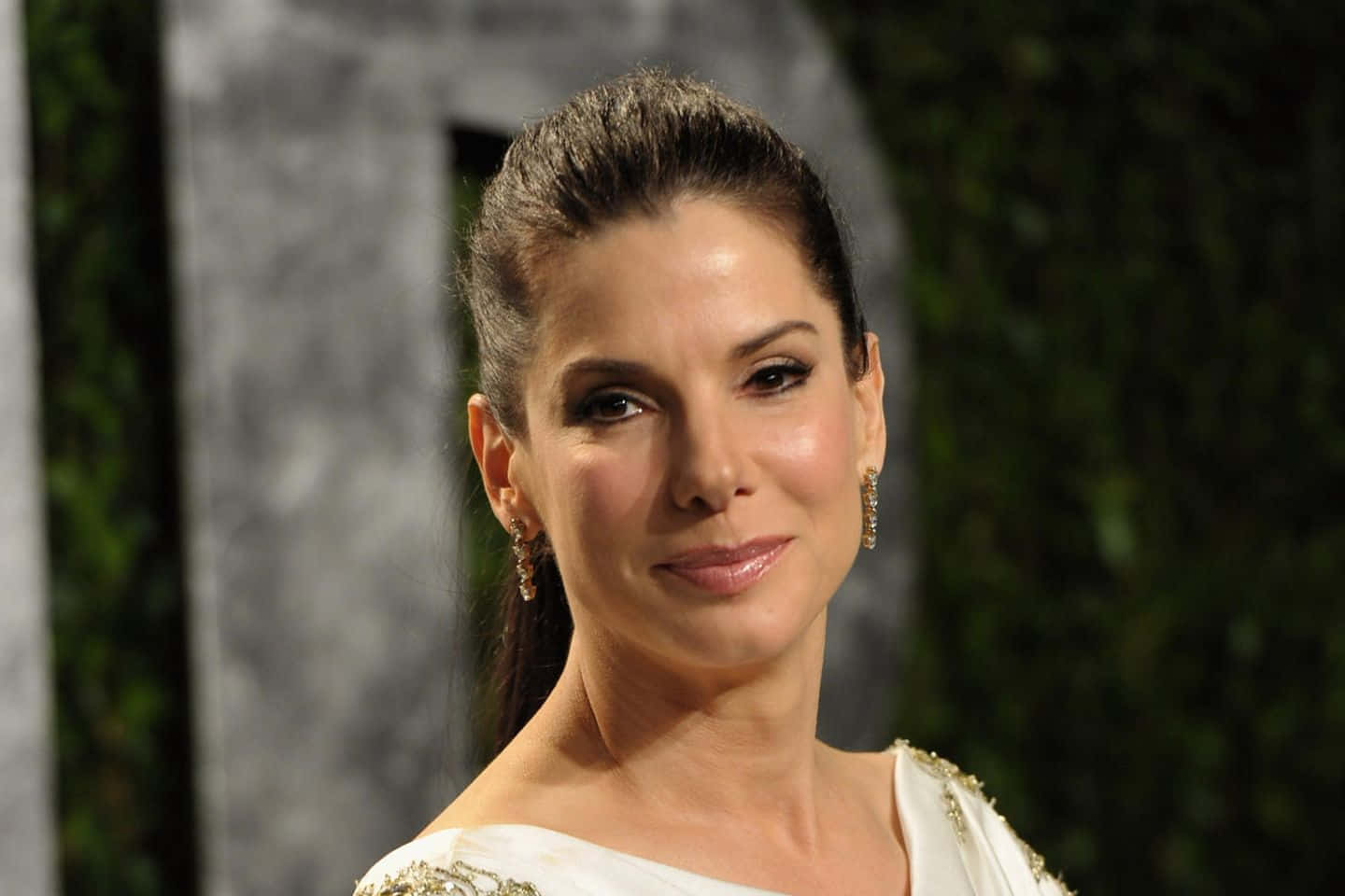 Sandra Bullock 2021 桌布 壁紙 — 一位深色頭髮向後梳的女性，穿著白色裝飾連衣裙，在模糊的綠色和灰色背景前輕柔微笑。