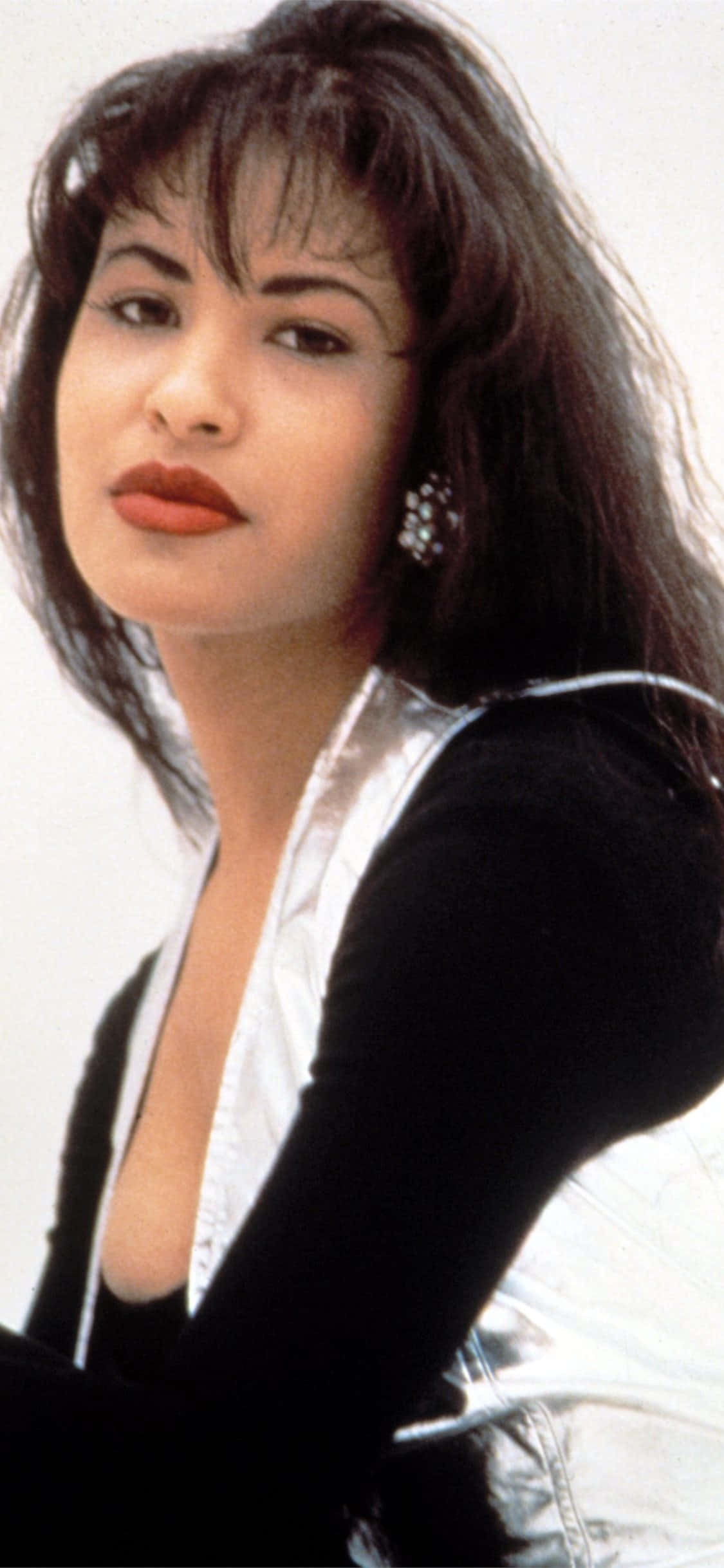 Selena Quintanilla iPhone 桌布 壁紙 — 一位深色頭髮和紅唇的女性，在純白色背景前穿著黑色和銀色的服裝。