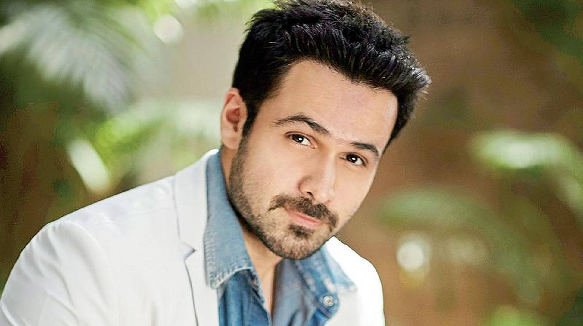Emraan Hashmi 桌布 壁紙 — 一位深色頭髮和鬍鬚的男人，在柔和的綠色樹葉背景前，穿著白色西裝外套內搭牛仔襯衫。