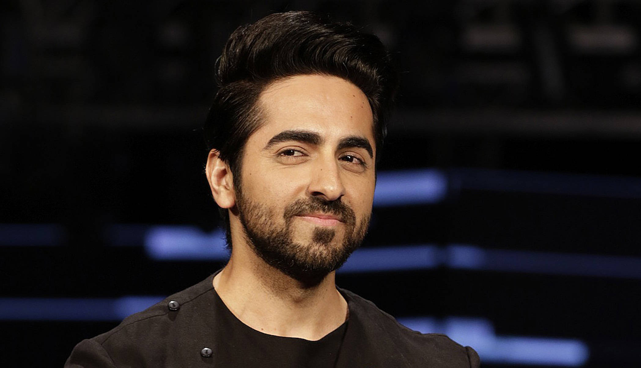 ayushmann khurrana 桌布 壁紙 — 一位深色頭髮和鬍鬚的男子，在帶有藍色燈光的模糊深色背景前溫柔微笑。