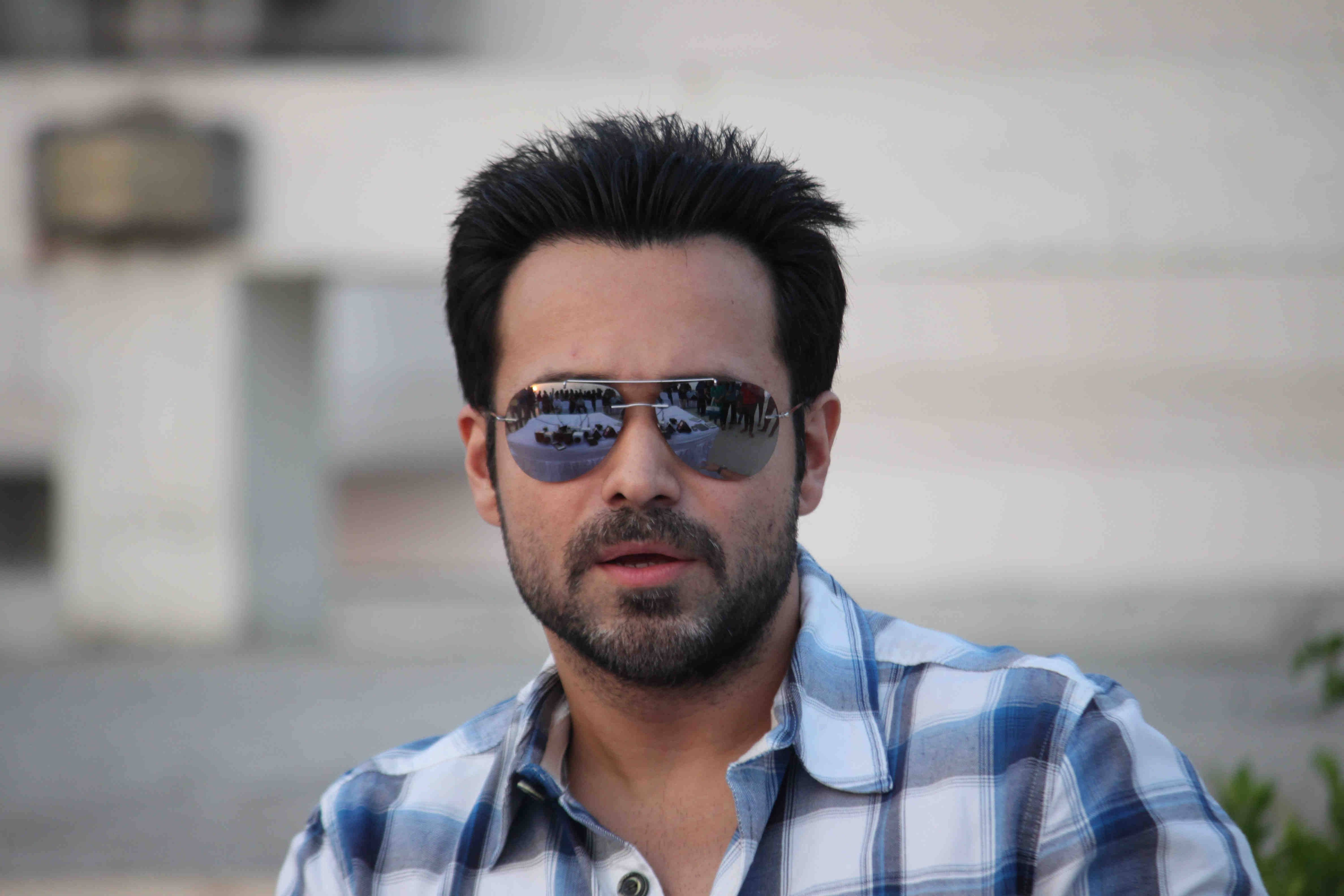 Emraan Hashmi 桌布 壁紙 — 一位深色頭髮和鬍鬚的男子，戴著反光飛行員太陽眼鏡和藍色格紋襯衫，背景是模糊的城市景觀。