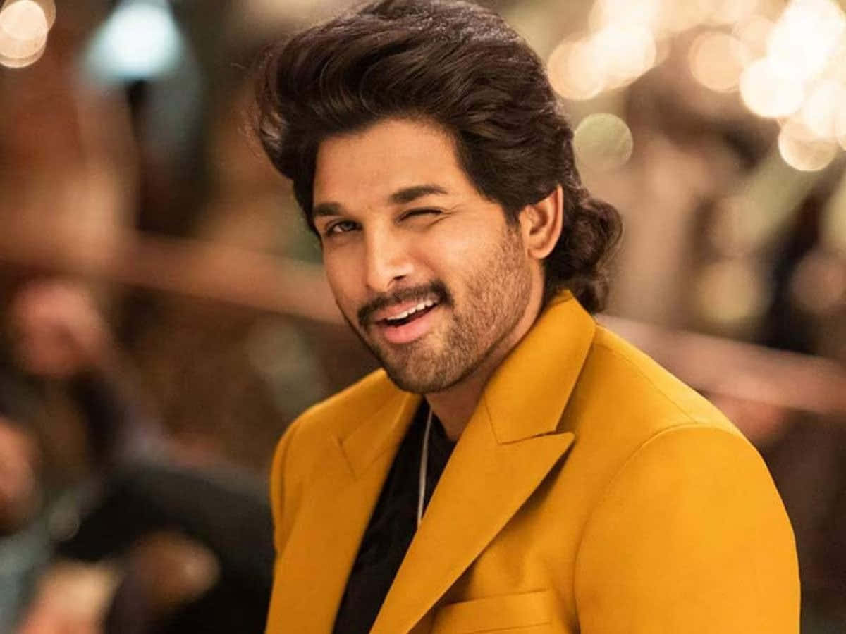 Allu Arjun 桌布 壁紙 — 一位深色頭髮和鬍鬚的男子，穿著亮黃色西裝外套，在柔和模糊的燈光背景下溫暖地微笑。