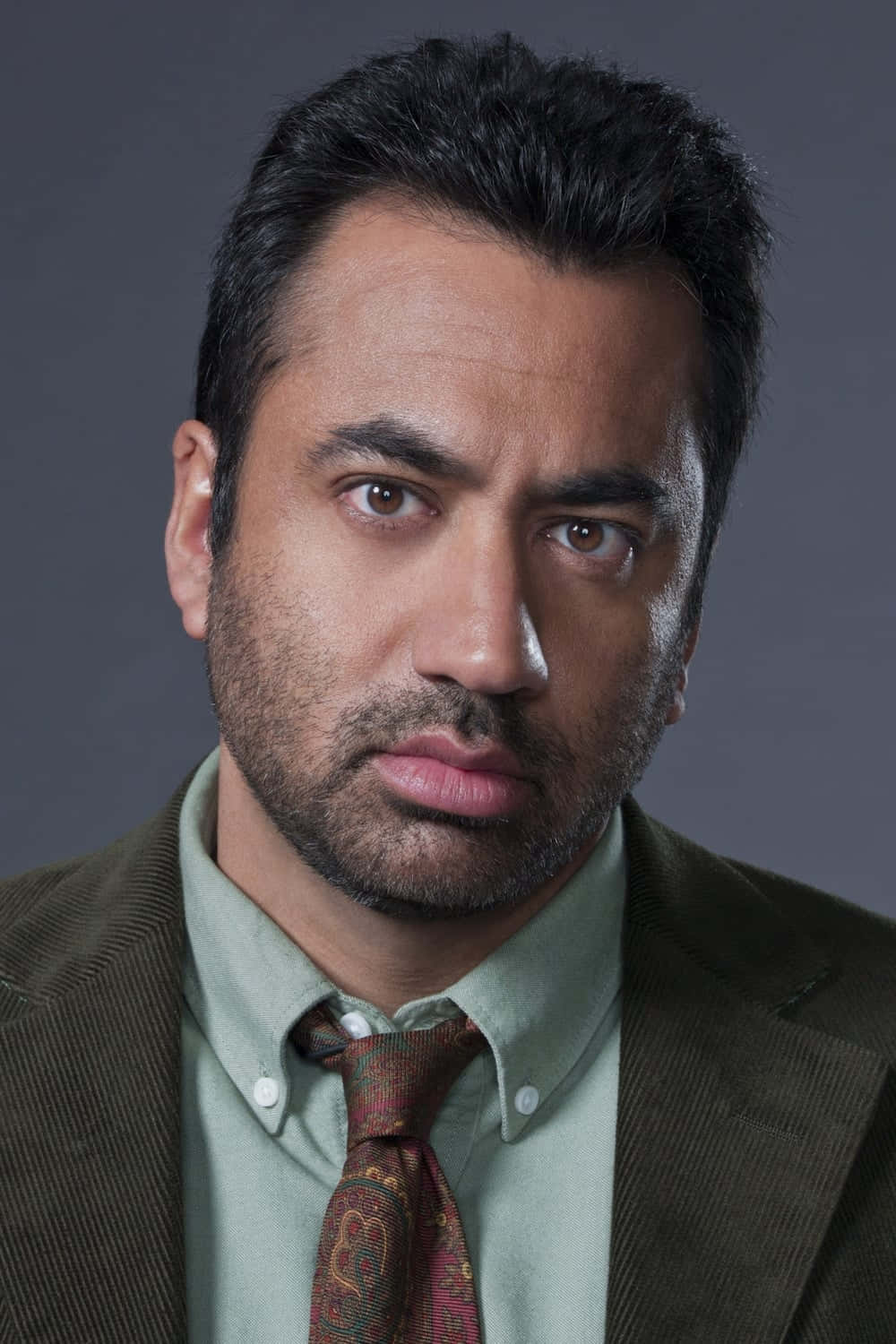 Kal Penn 桌布 壁紙 — 一位深色頭髮和鬍鬚的男子，穿著棕色西裝和圖案領帶，背景為灰色。