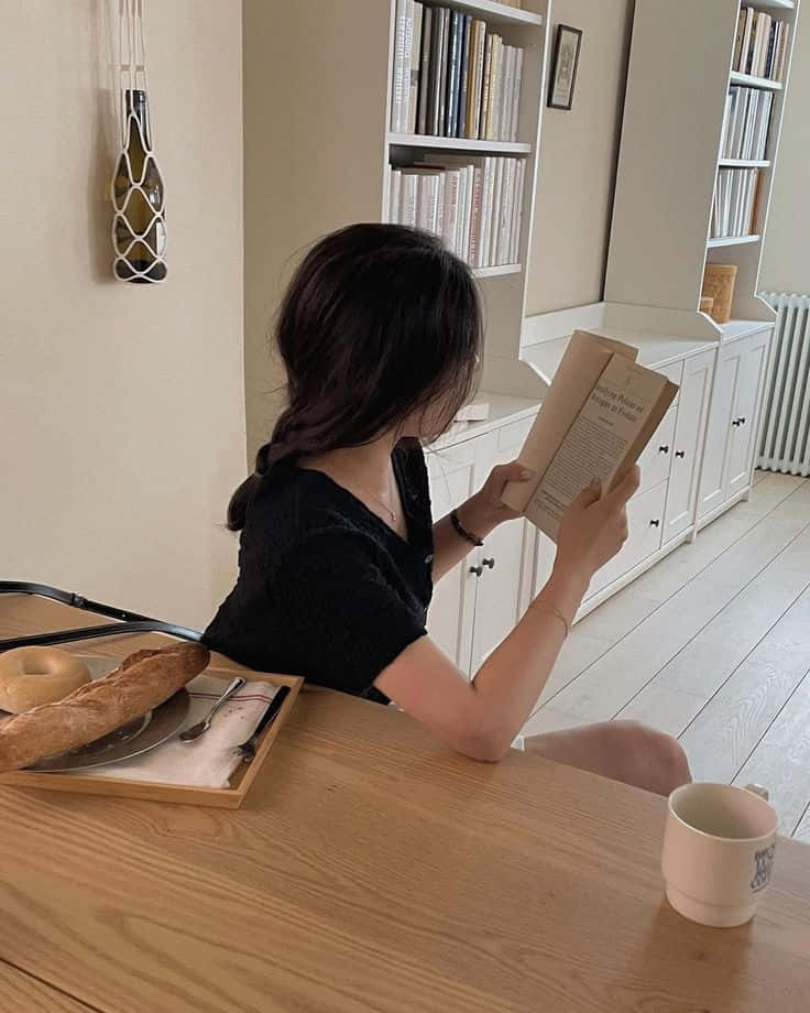 美學米色 桌布 壁紙 — 一位深色頭髮的人坐在木桌旁閱讀一本書，早餐物品放在附近，房間明亮，有白色書架。