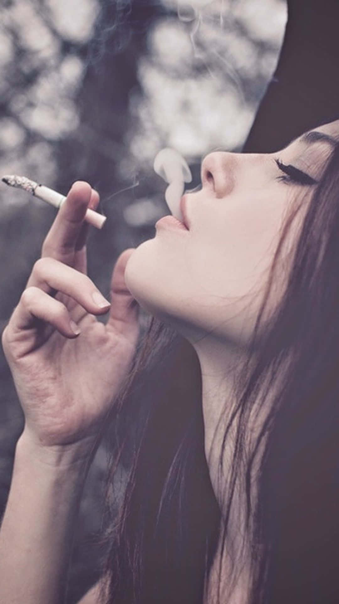 Girl Smoking 桌布 壁紙 — 一位深色頭髮的女性在戶外吸煙，煙霧從她的嘴唇繚繞，背景是模糊的自然景色。