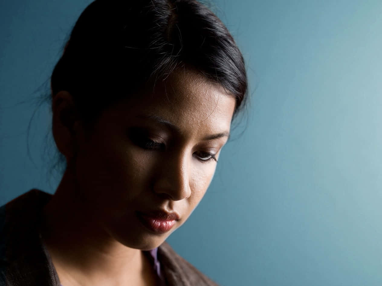 Depression Girl 桌布 壁紙 — 一位深色頭髮的女性在柔和而戲劇性的燈光下，對著柔和的藍色背景向下凝視。