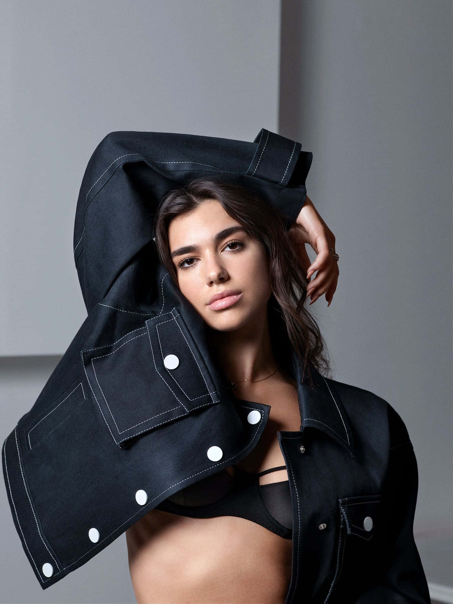 Dua Lipa 桌布 壁紙 — 一位深色頭髮的女性在極簡灰色背景前，將黑色牛仔外套舉過頭頂。