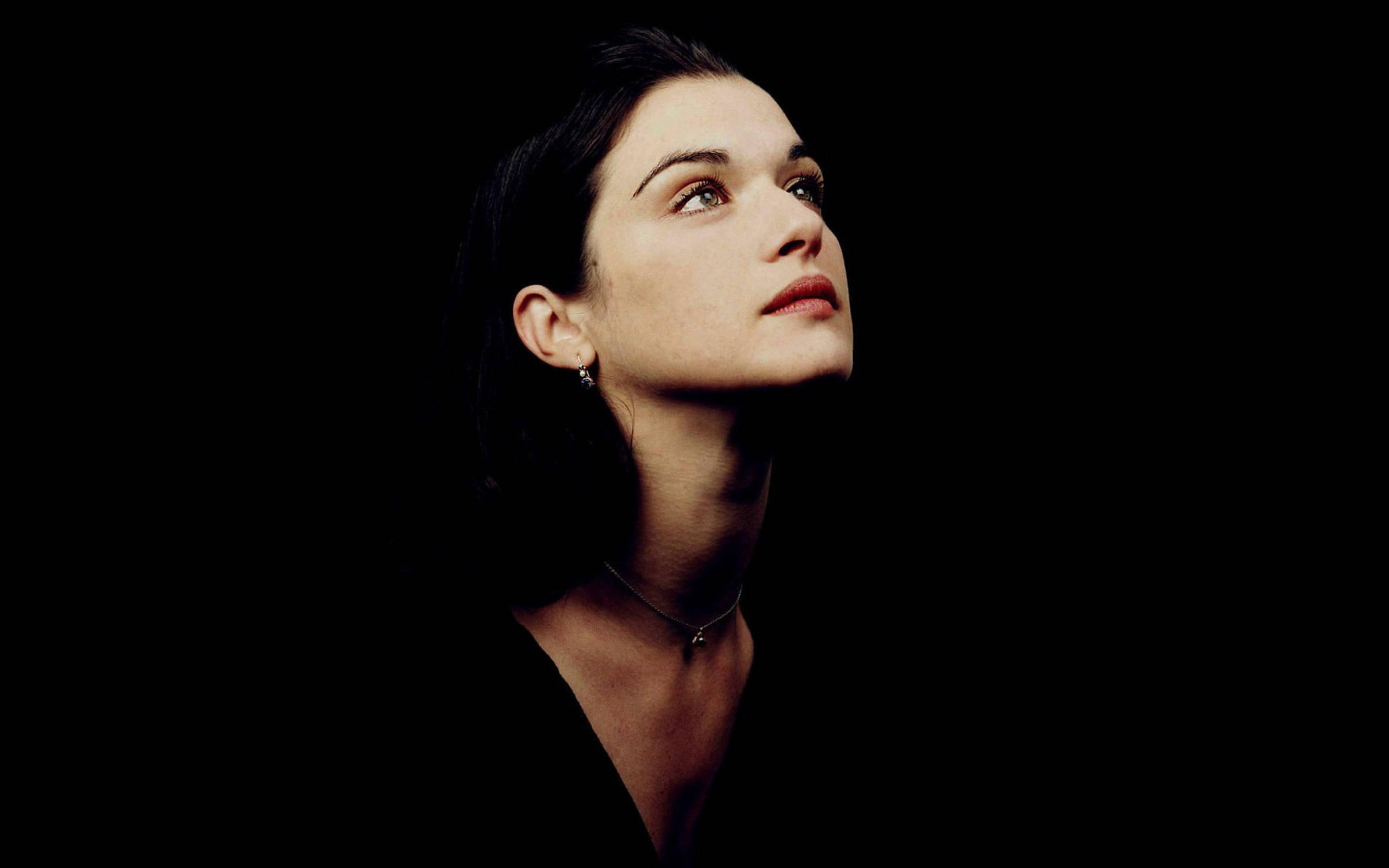 Rachel Weisz 桌布 壁紙 — 一位深色頭髮的女性在純黑背景下向上凝視，她的臉部被柔和地照亮，突顯出她沉思的表情。