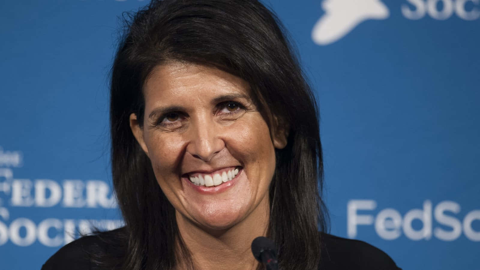 Smile 桌布 壁紙 — 一位深色頭髮的女性在藍色FedSoc背景前的新聞活動中燦爛微笑。
